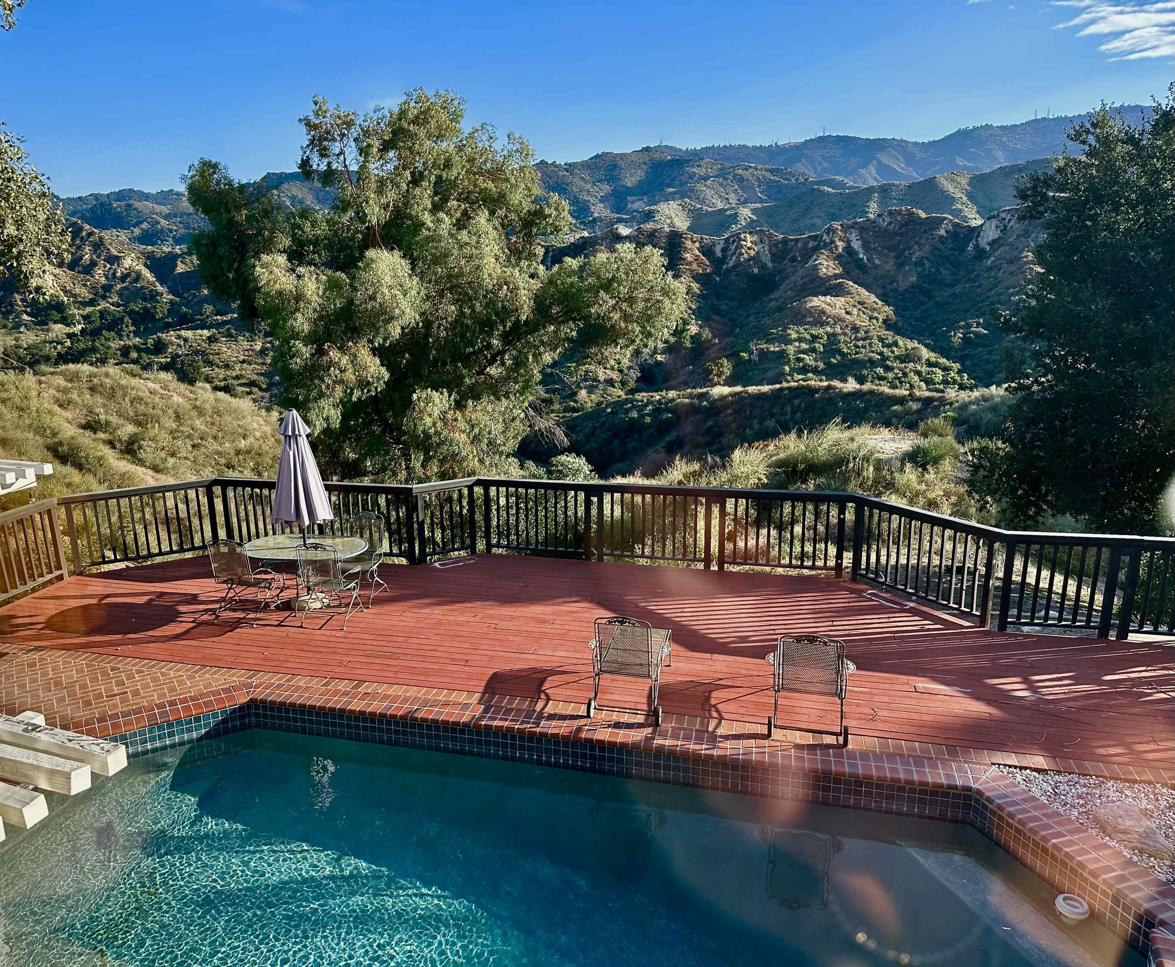 A deck with a swimming pool overlooks a hilly landscape under a clear sky.