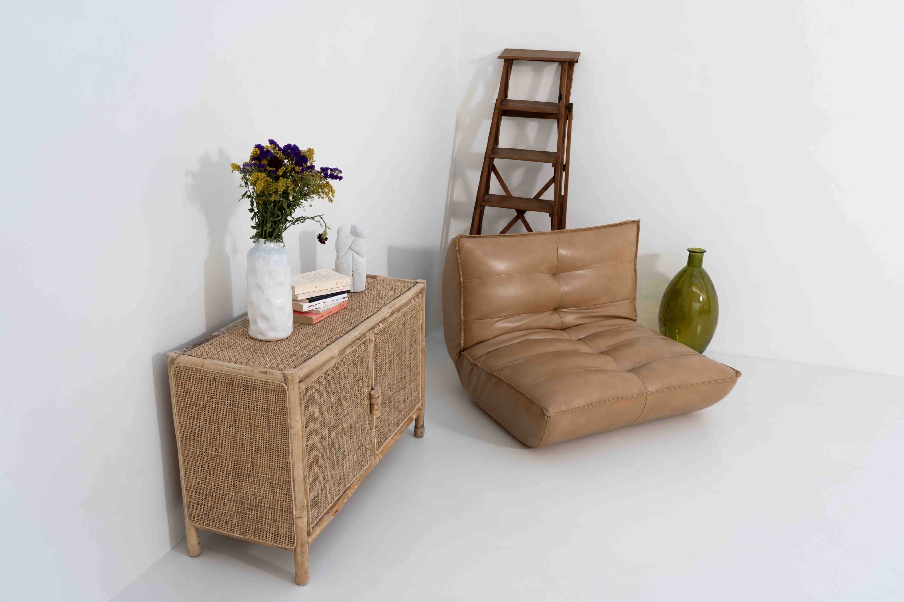The image shows a minimalist interior setting featuring a beige leather chair, a wicker cabinet with a flower vase, and a wooden ladder against a white wall.