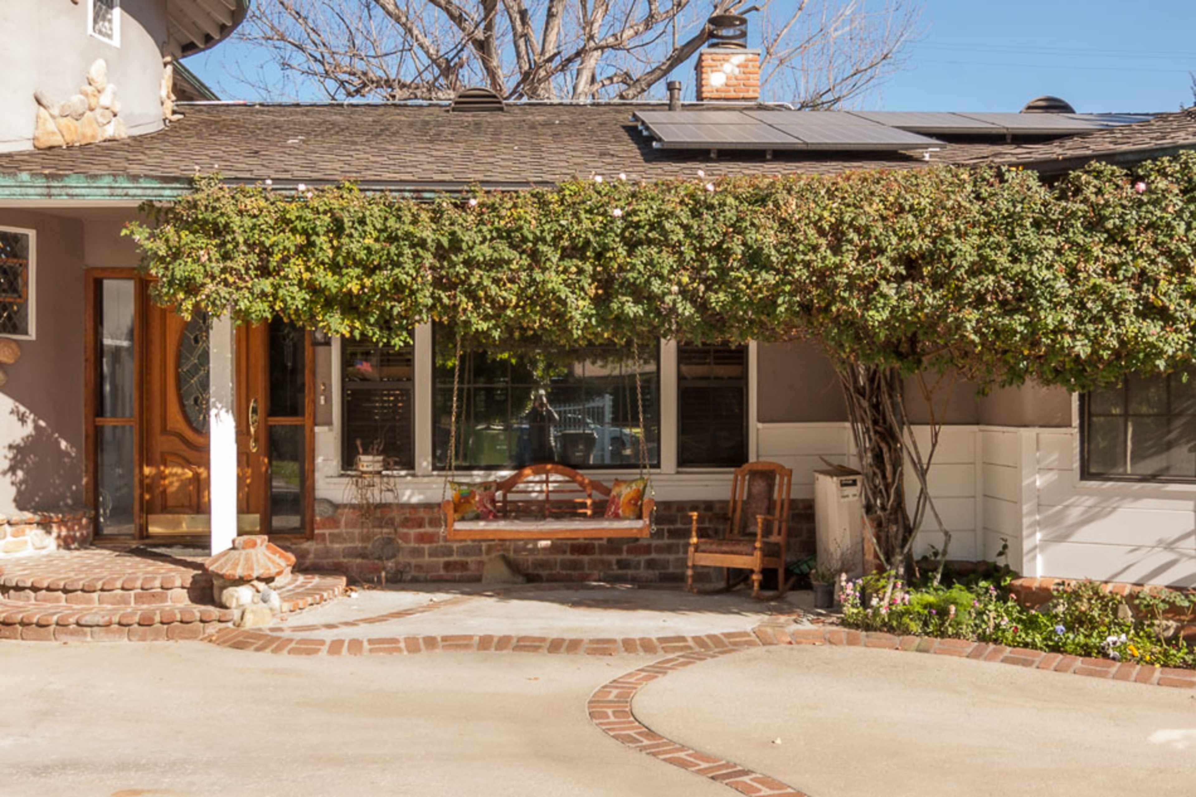 A house with a large window and a covered porch, surrounded by greenery and a brick pathway leading to the entrance.