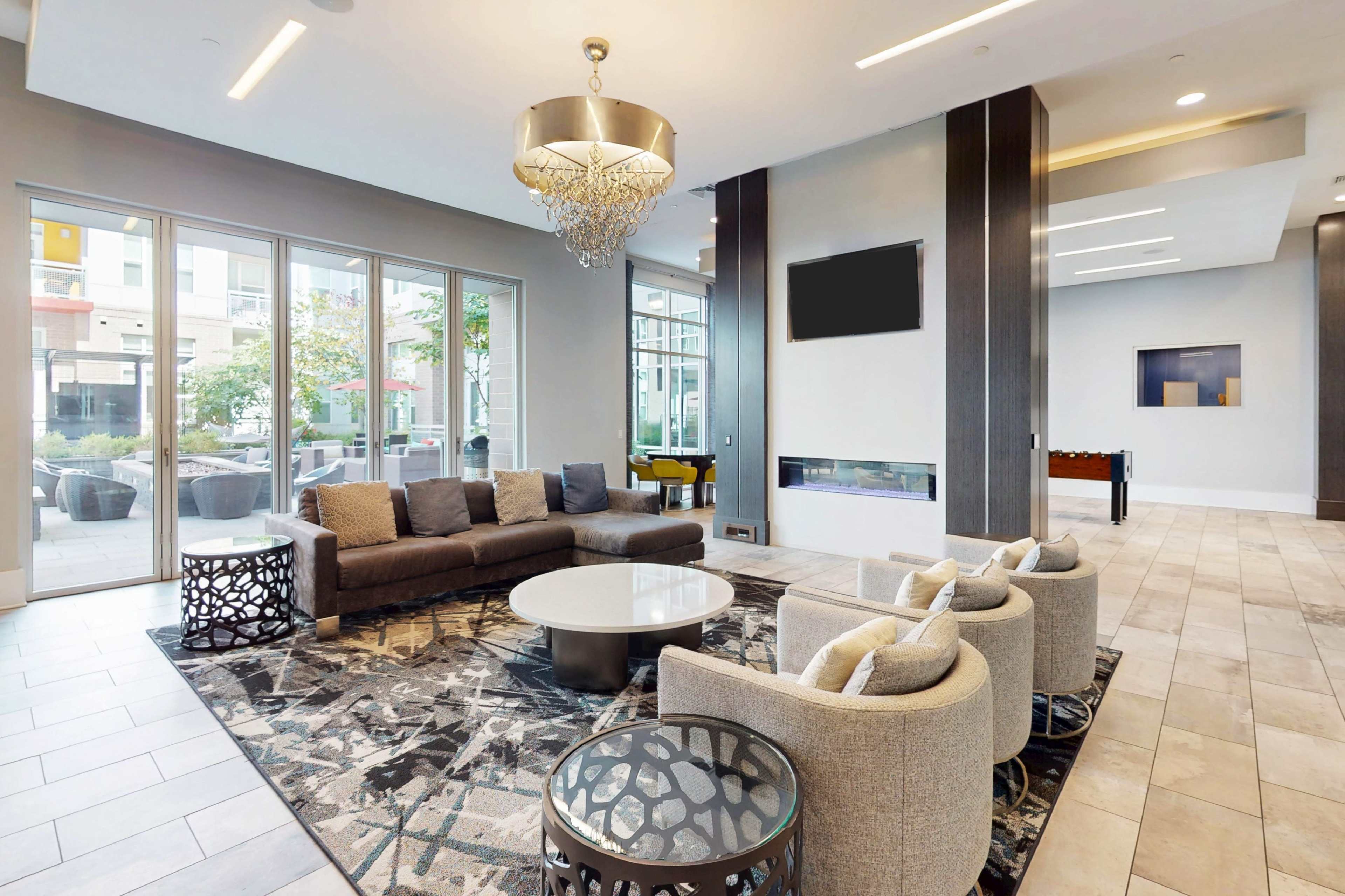 The image shows a modern lounge area featuring a sectional sofa, circular coffee table, and two additional chairs, all arranged around a patterned rug, with large windows letting in natural light.
