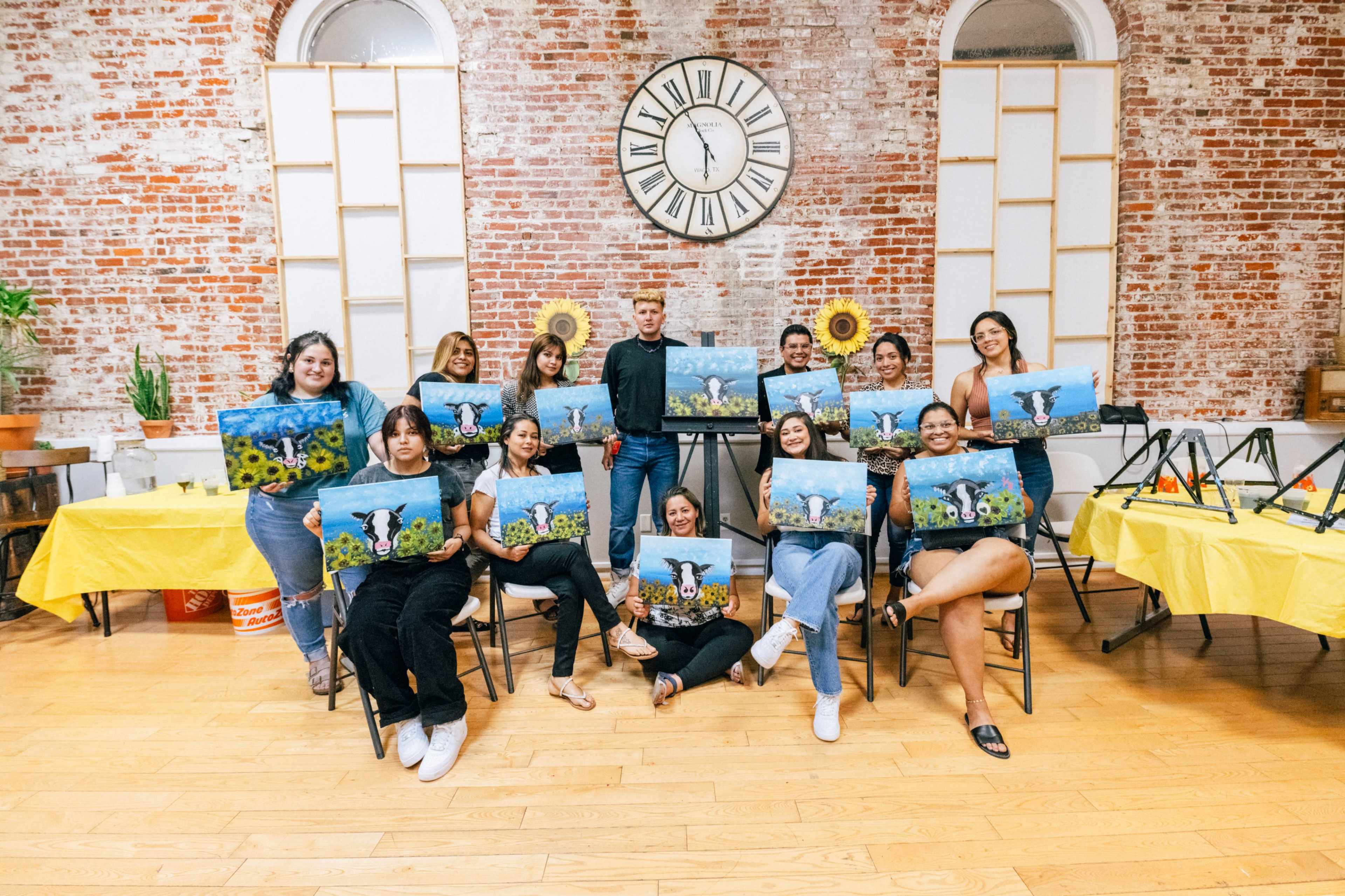 A group of people is posing indoors with paintings they created, surrounded by brick walls and a large clock, while seated at tables covered with yellow cloths.
