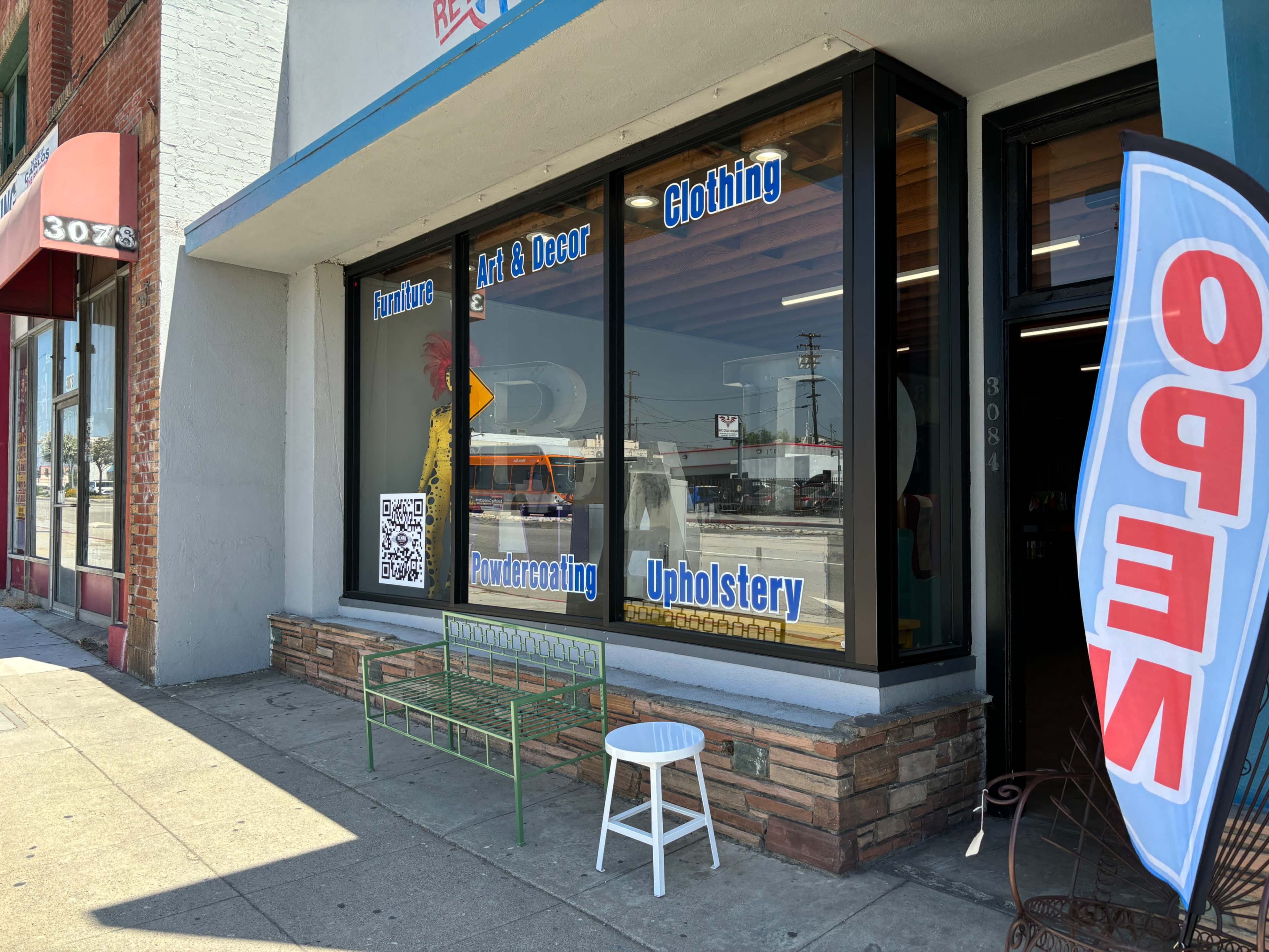 A storefront with large windows displaying signs for furniture, art, decor, clothing, powder coating, and upholstery, alongside a bench and a stool outside.
