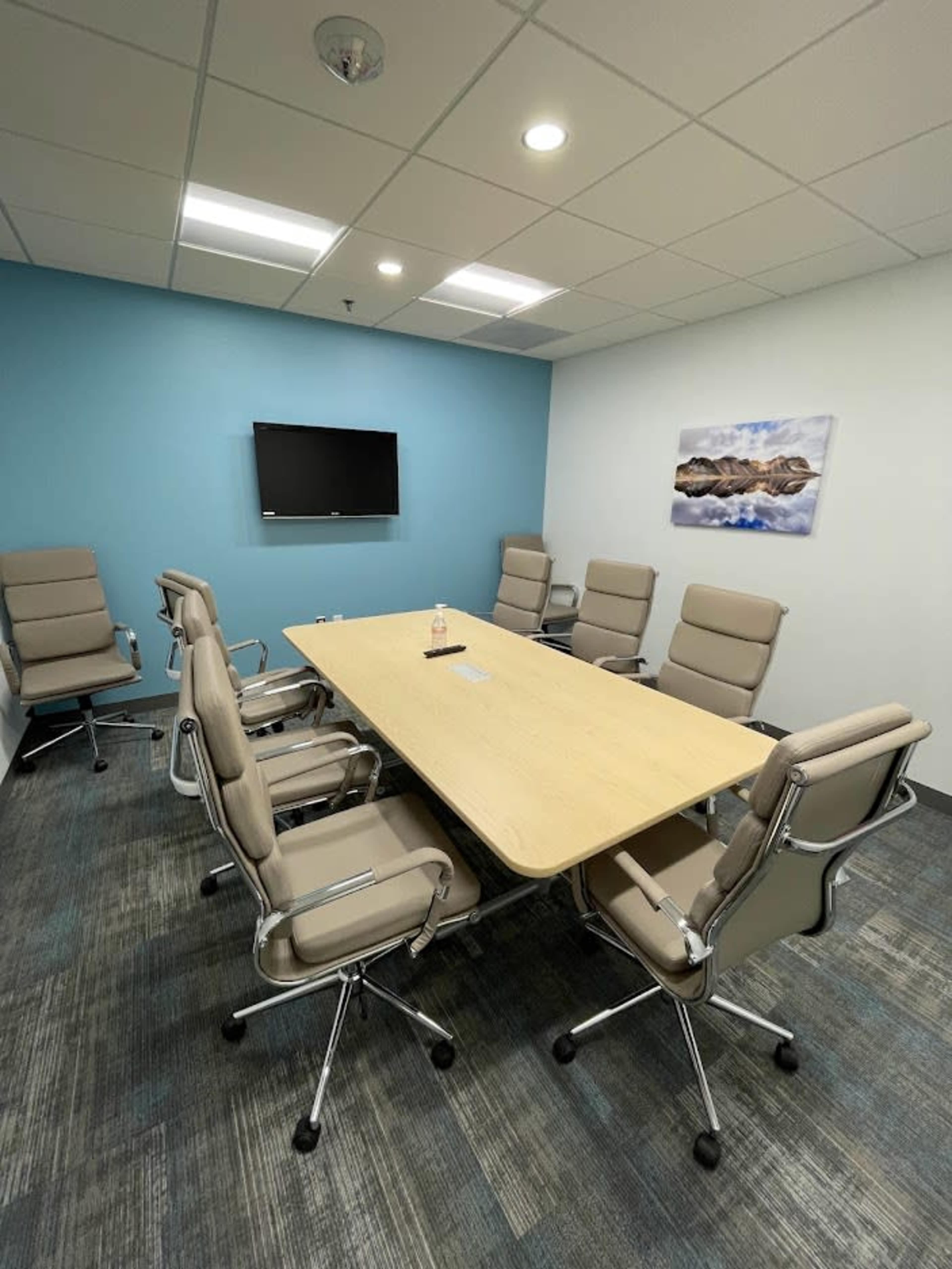 A conference room features a long wooden table surrounded by six beige chairs, with a television mounted on a blue wall and a landscape print hanging nearby.