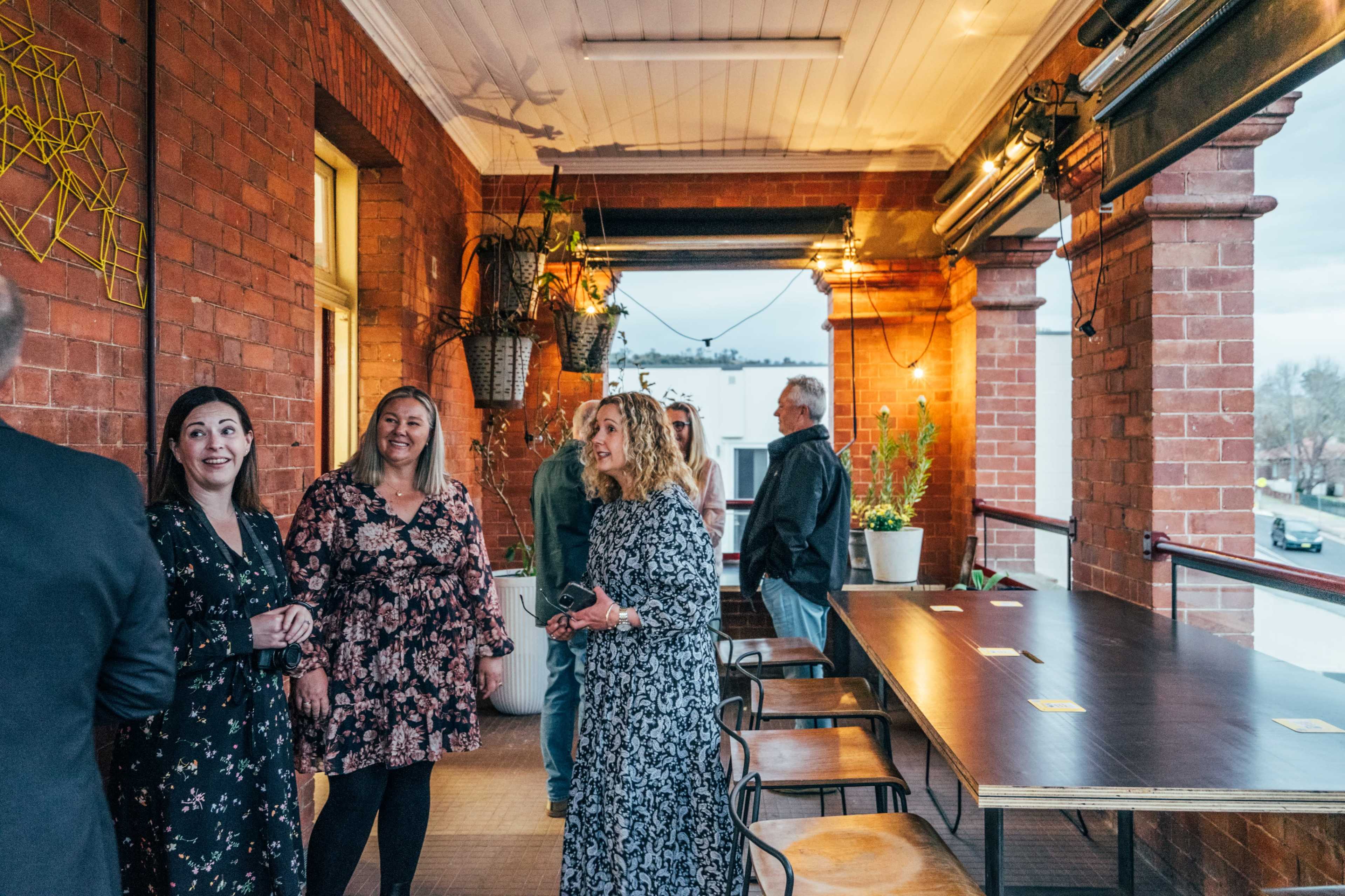 A group of people is standing and conversing on a balcony with brick walls and potted plants in the background.