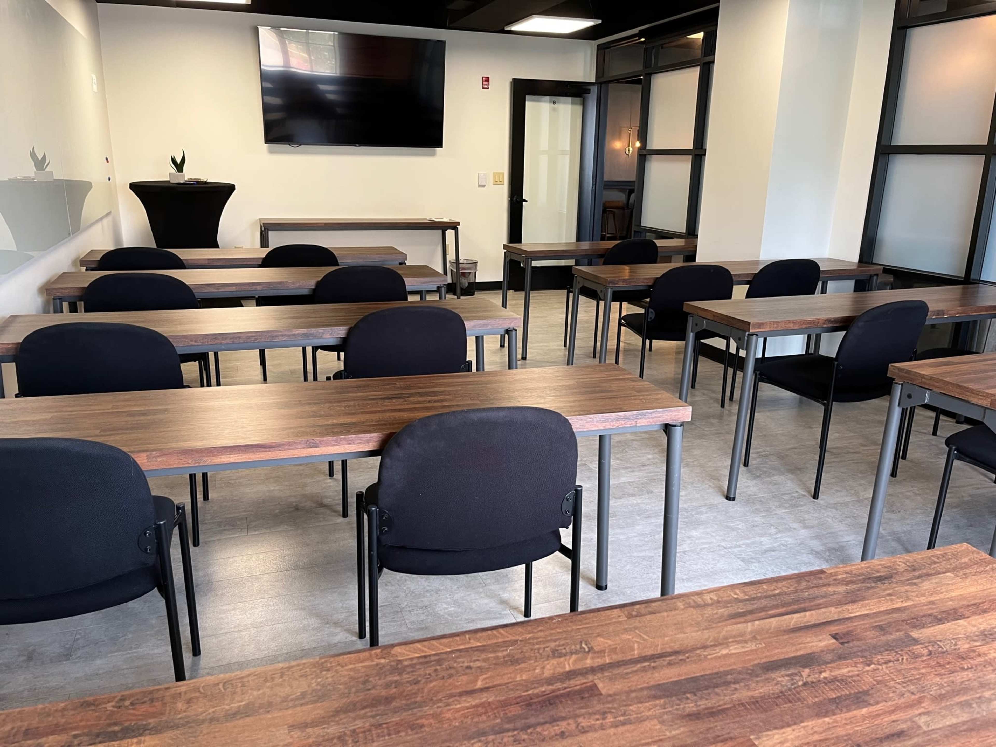 A classroom-style setup with several rows of wooden tables and black chairs arranged in a modern, well-lit space featuring a television screen on the wall.