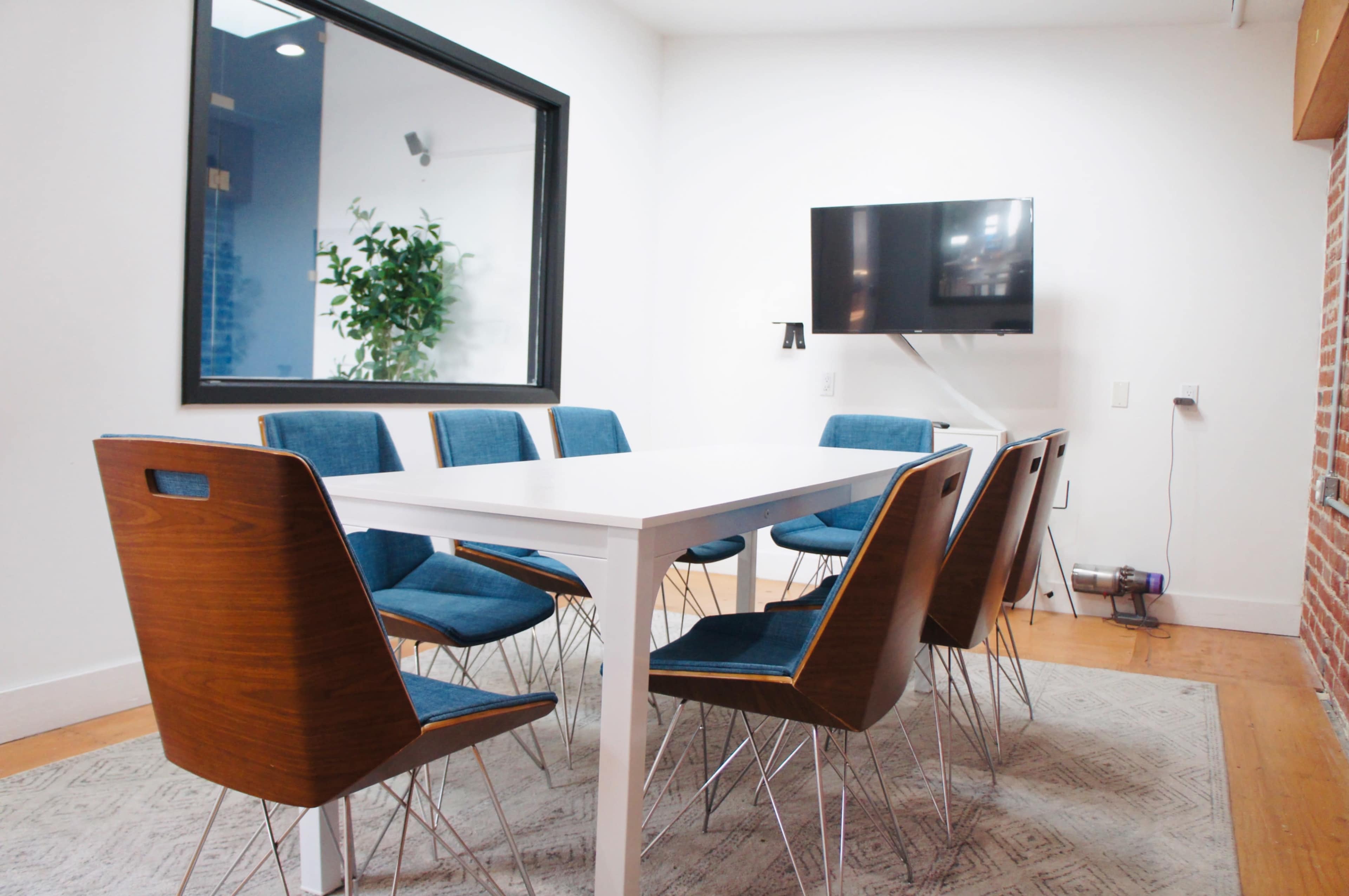 A modern conference room features a white table surrounded by eight wooden chairs with blue upholstery, a large window, and a wall-mounted television.