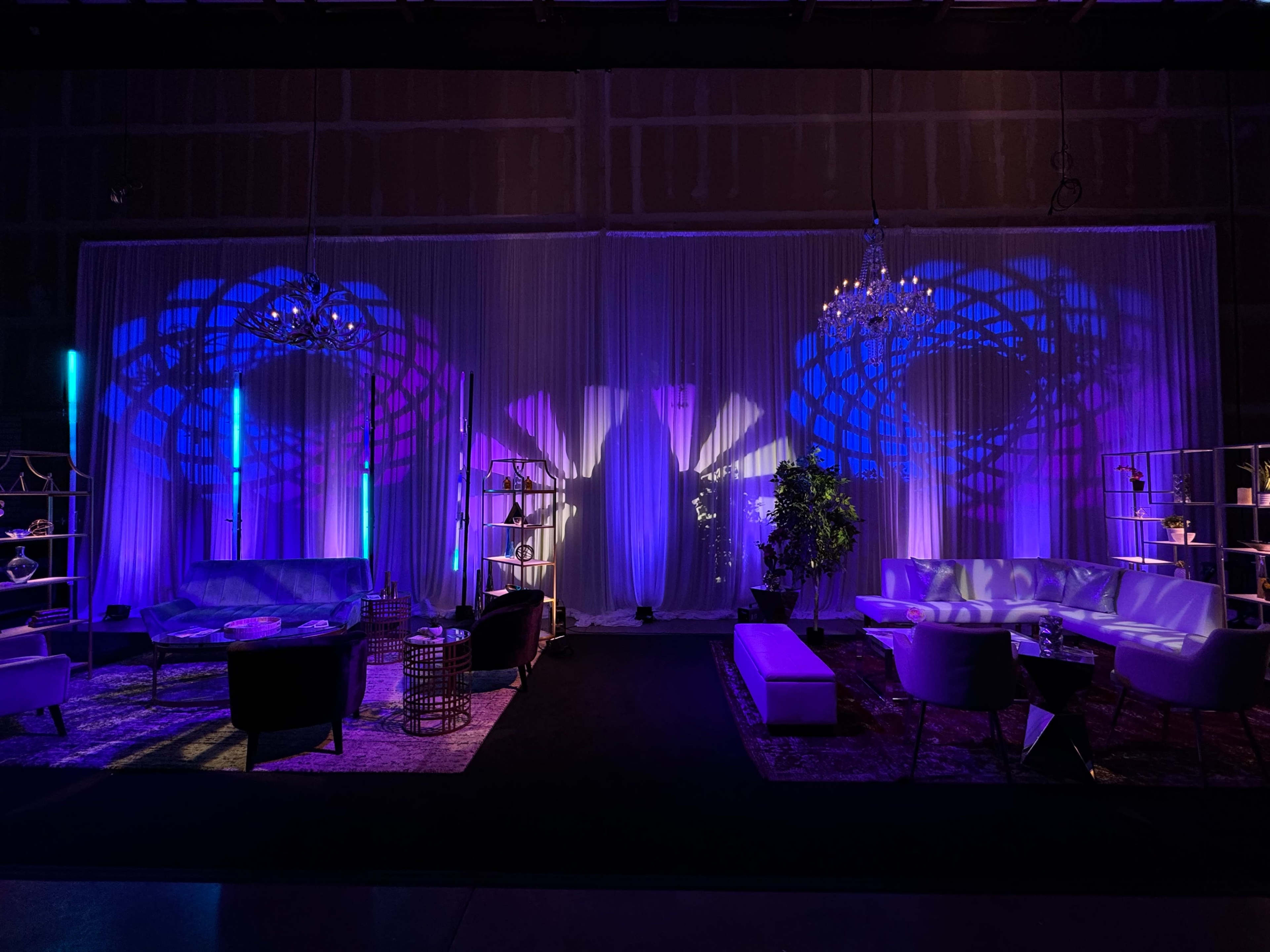 A stylish event space with a black carpet, white drapes, and purple lighting illuminating the walls, creating intricate shadow patterns behind modern seating arrangements.