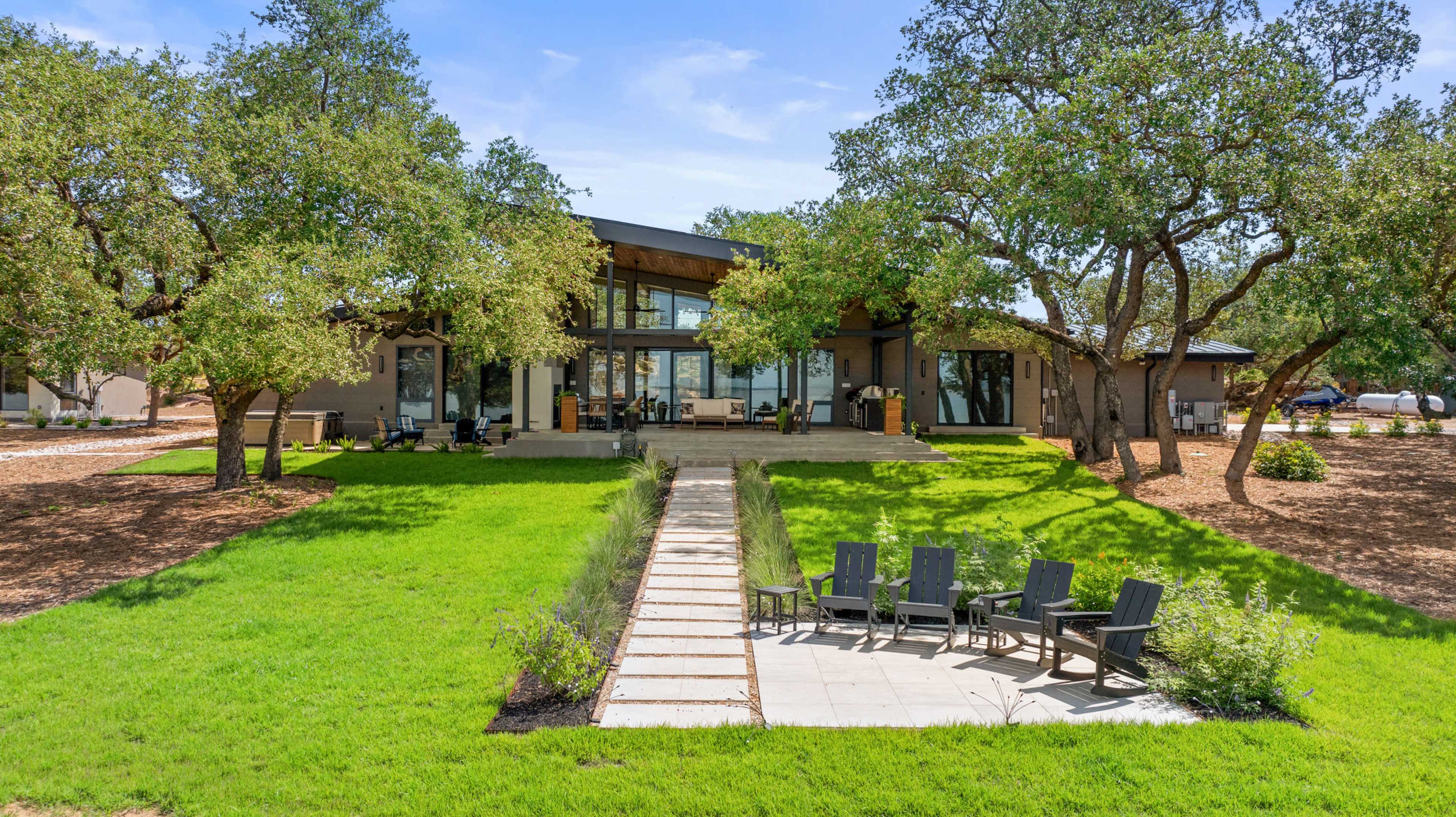 The image shows a modern home surrounded by lush green grass, with a stone pathway leading to a cluster of outdoor chairs under large trees.