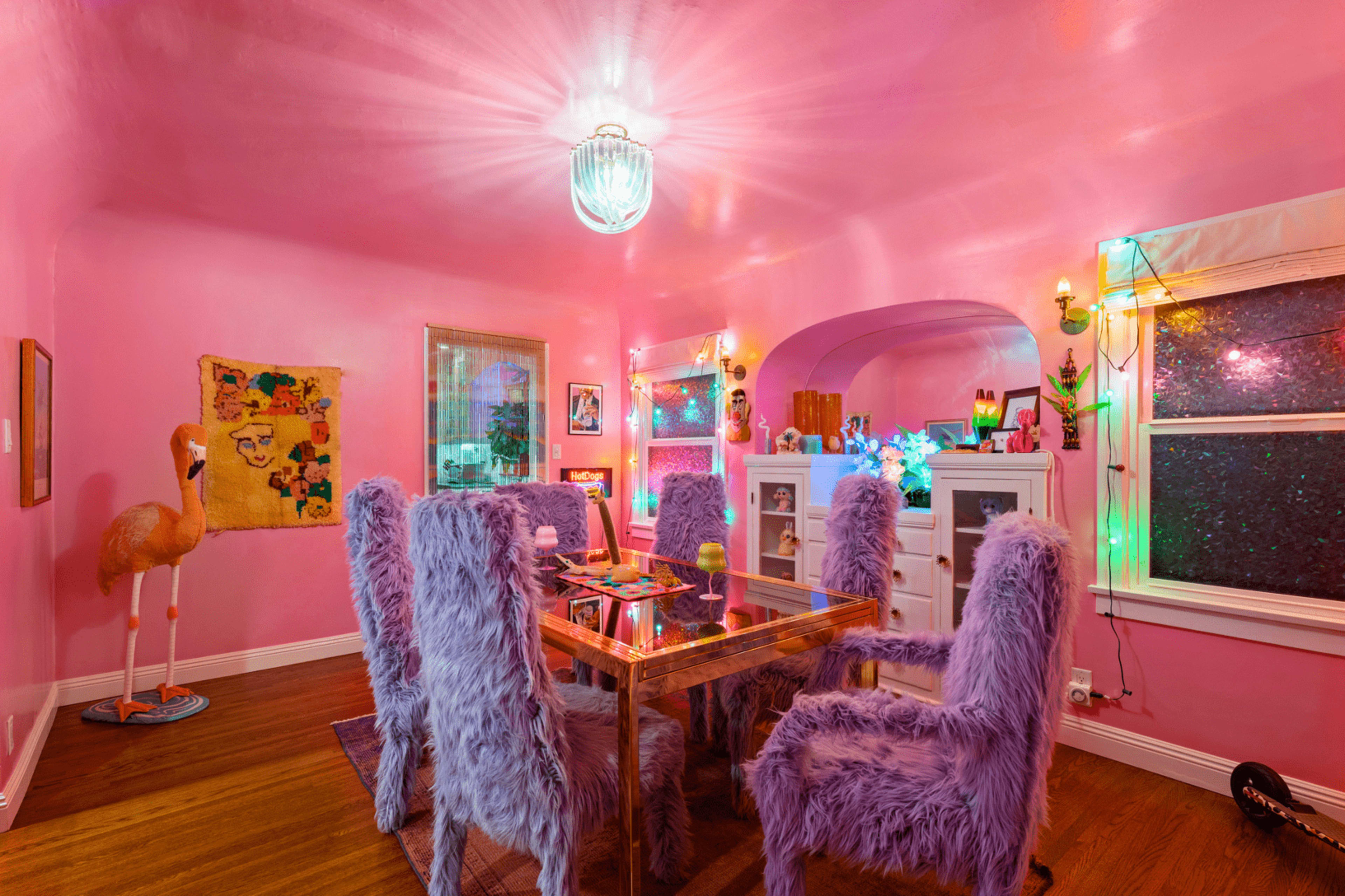 A dining room features a glass table surrounded by purple, fur-covered chairs, bright pink walls, and colorful decorations with a flamingo statue in one corner.