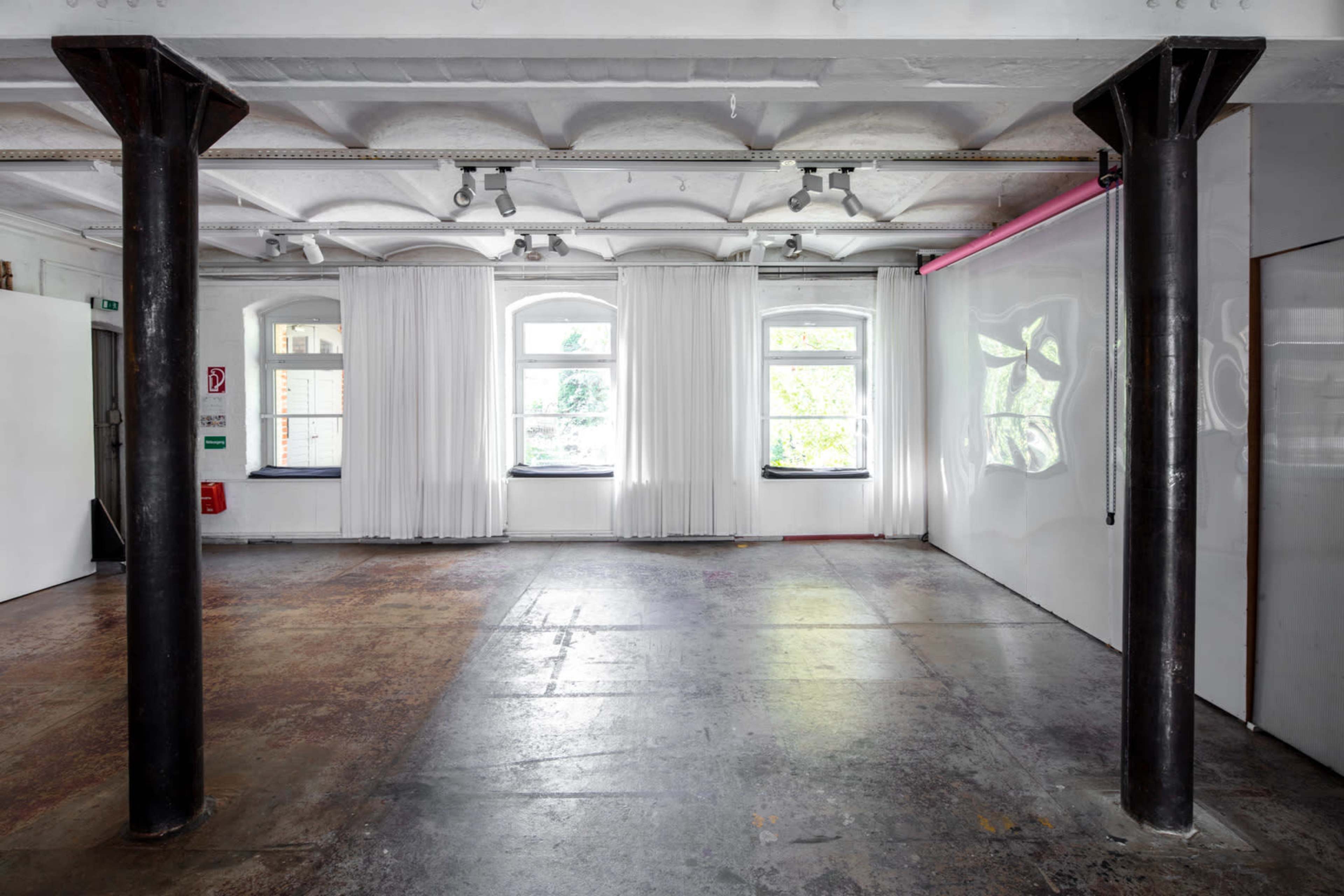 The image shows a spacious, bright room with white curtains, large windows, and two support columns.