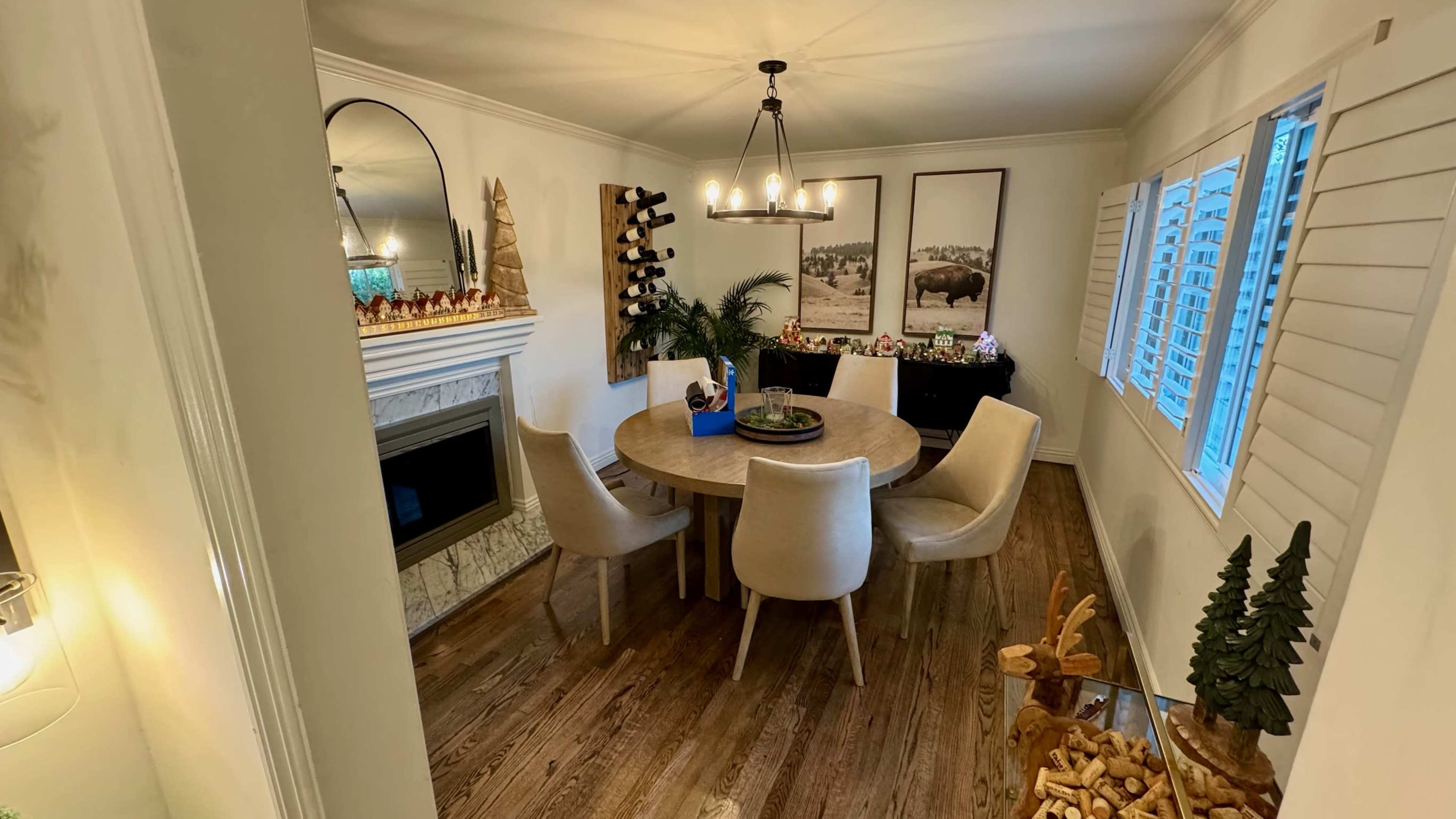 The image shows a dining area featuring a round wooden table surrounded by upholstered chairs, with a fireplace and decorative elements in the background.