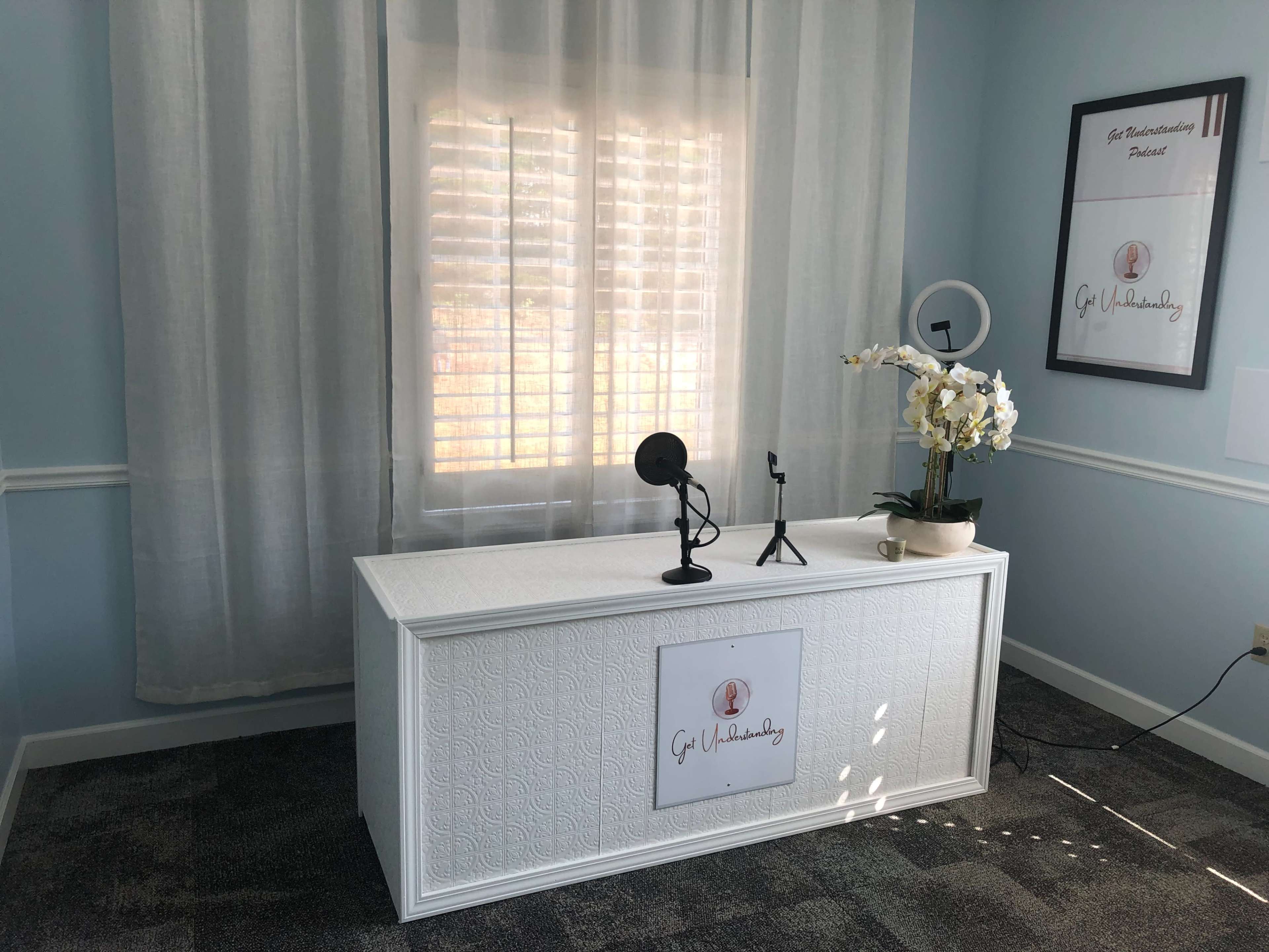 The image shows a brightly lit room with a white desk featuring a microphone, a lamp, and an ornamental plant, situated near a window with plantation shutters.