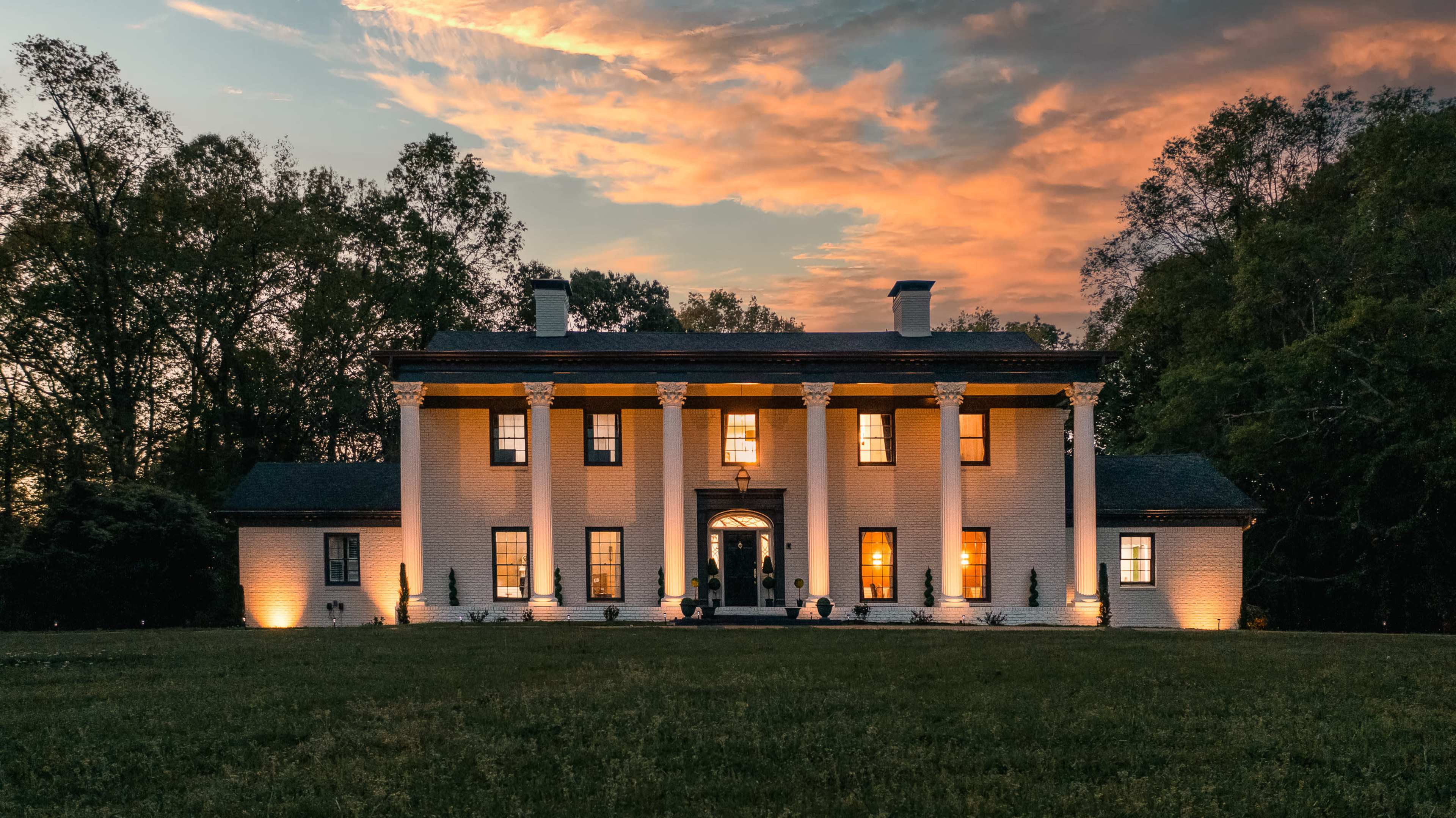 A large, two-story white brick mansion with columns is set against a colorful sunset and surrounded by trees.