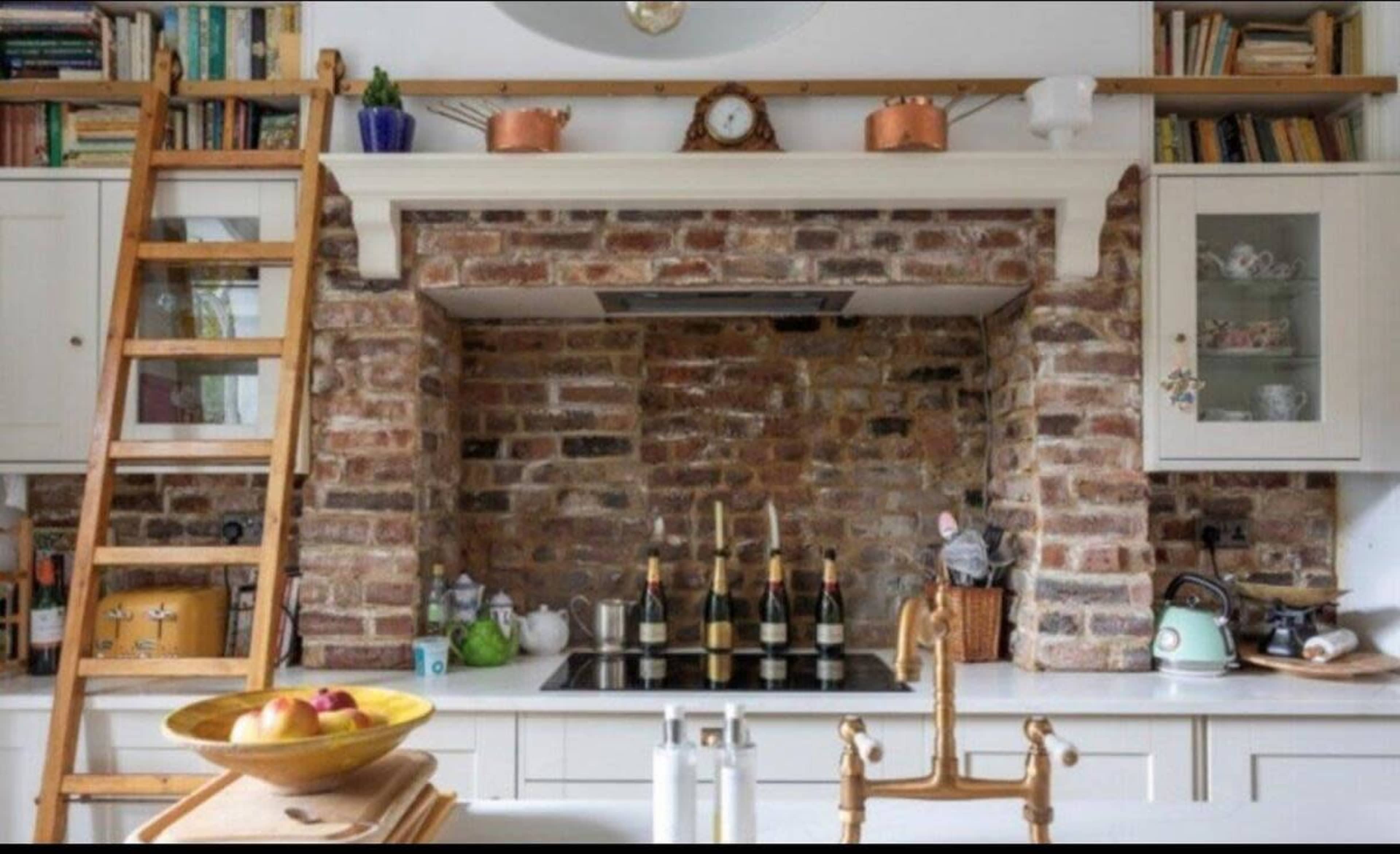 A rustic kitchen with a brick feature wall, a stove beneath a brick arch, and wooden shelves holding various kitchen items.