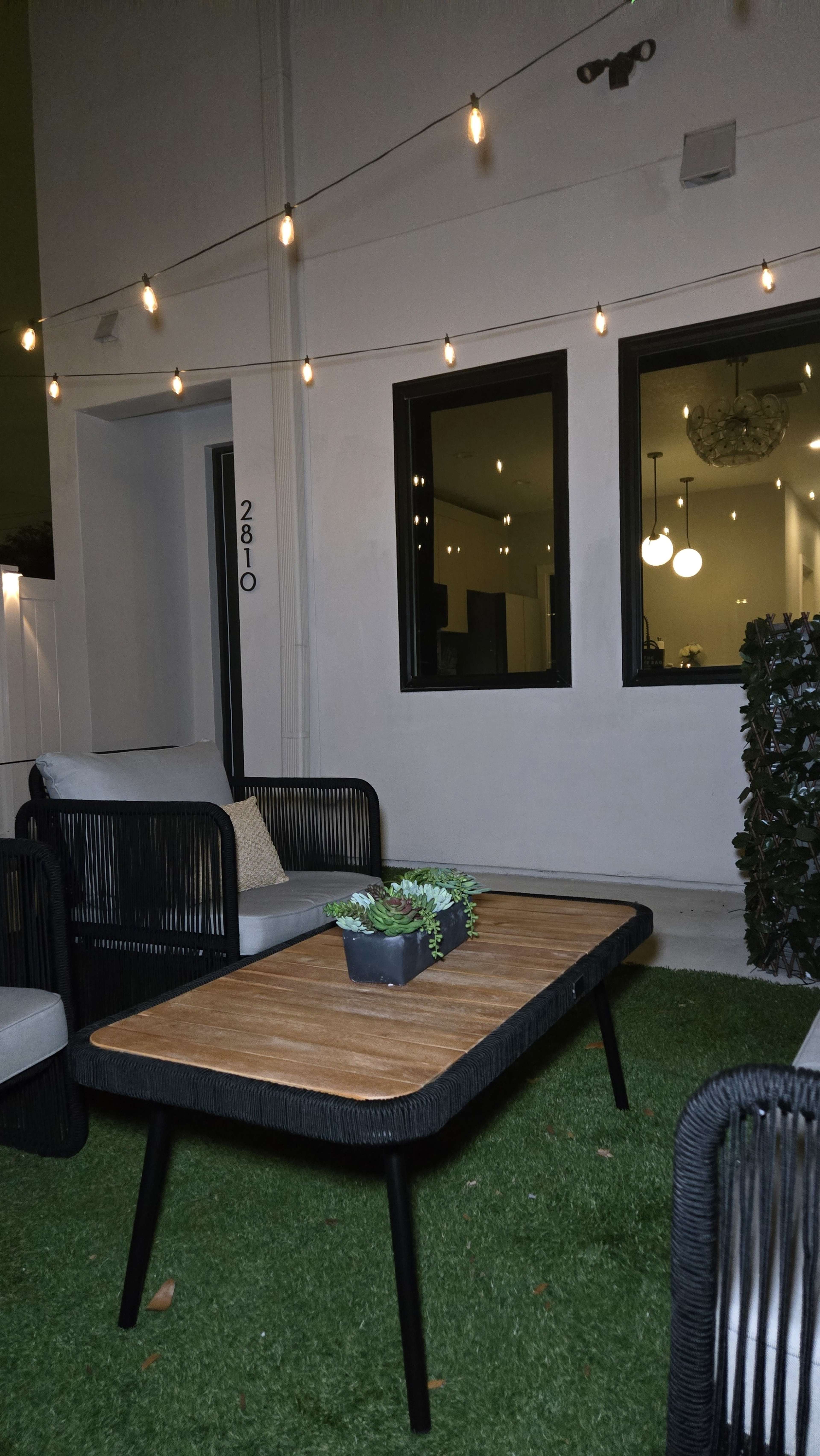 The image shows a modern outdoor seating area with two chairs, a wooden table, and string lights above, set on a grass-like surface.