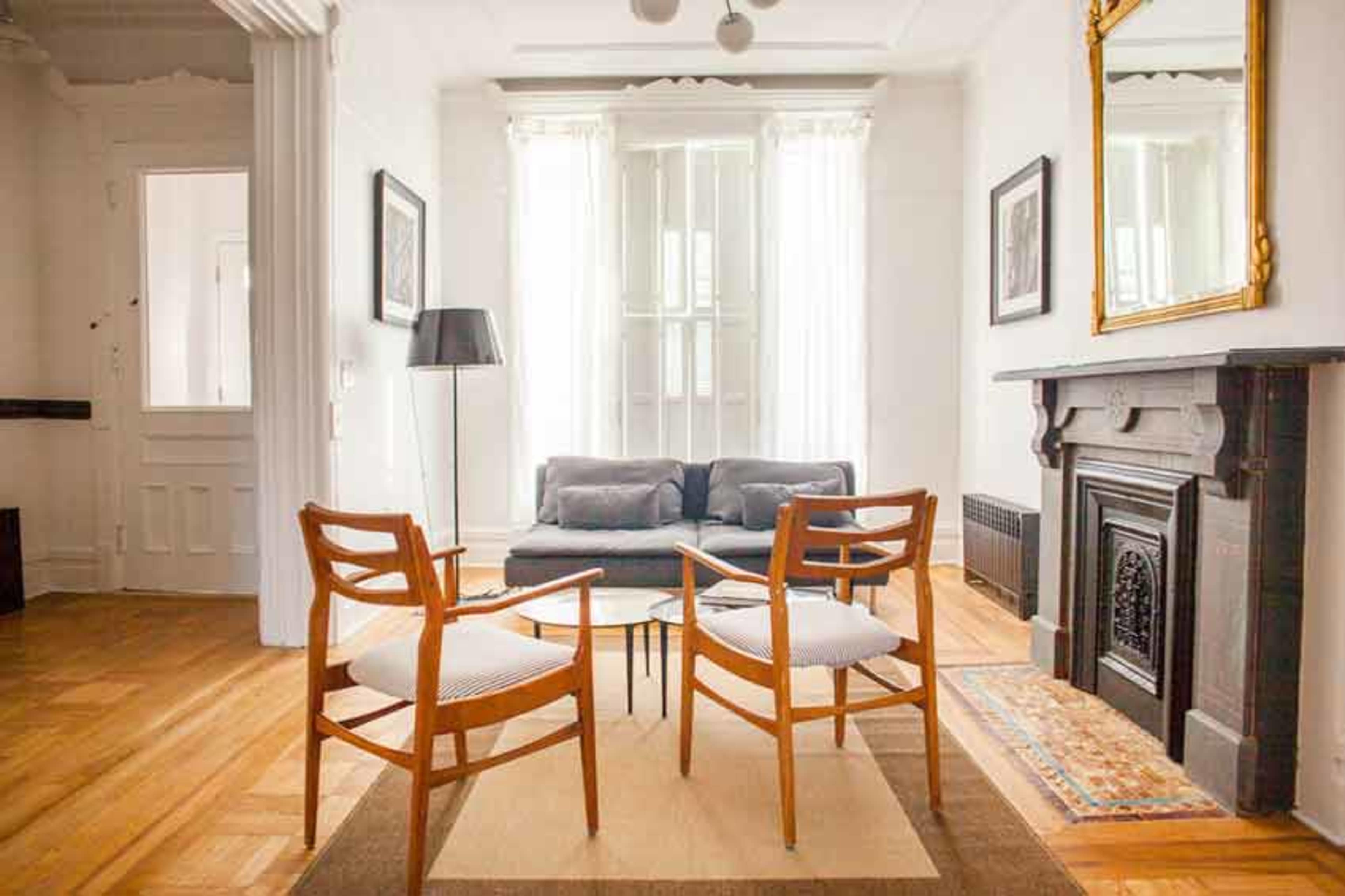 The image shows a bright living room featuring two wooden chairs facing a coffee table, a gray sofa, and a fireplace with a decorative mirror above it.