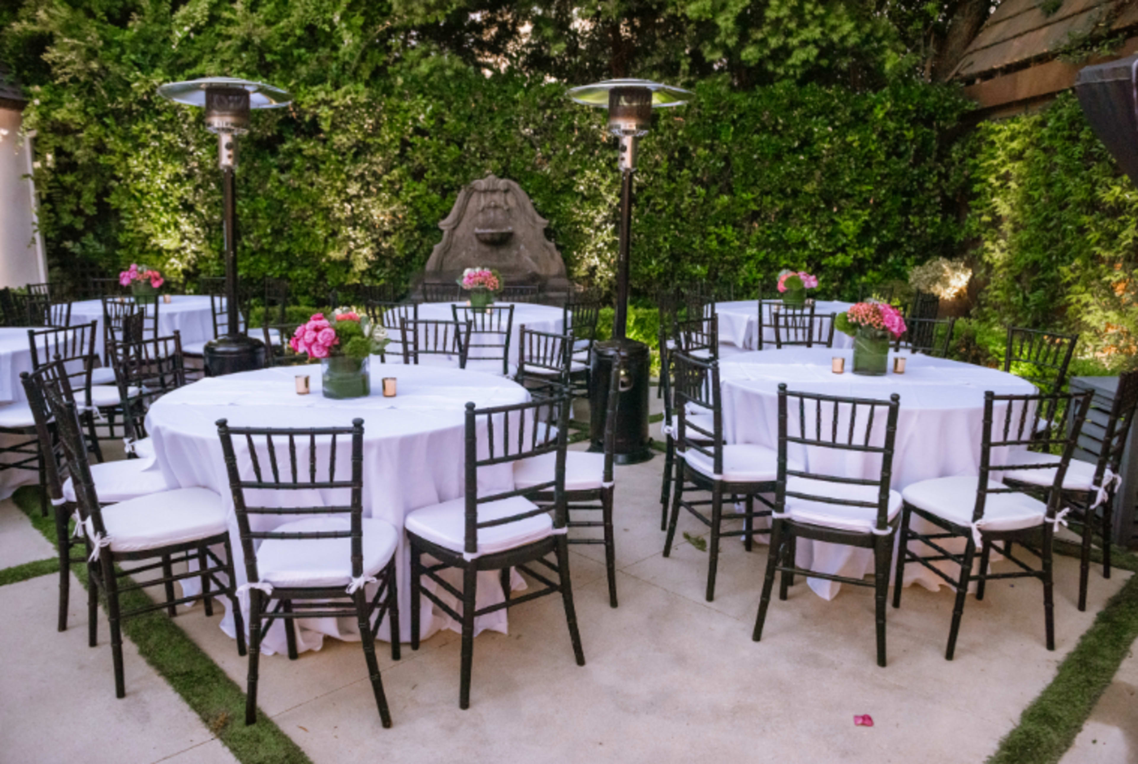 The image shows a patio area set with several round tables covered in white tablecloths, surrounded by black chairs and adorned with pink flowers in the center.