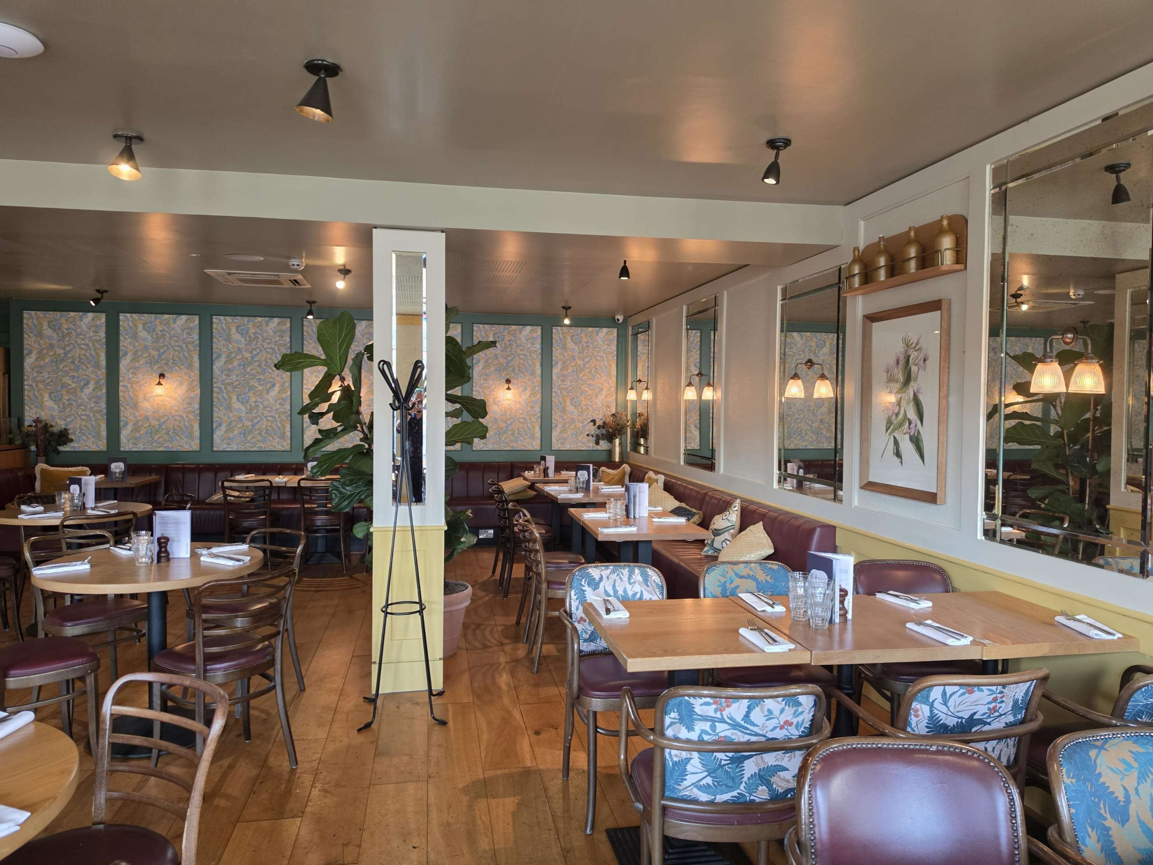 The image shows an indoor restaurant setting with wooden tables, stylish chairs, and decorative plants, featuring a patterned wall and mirrors.