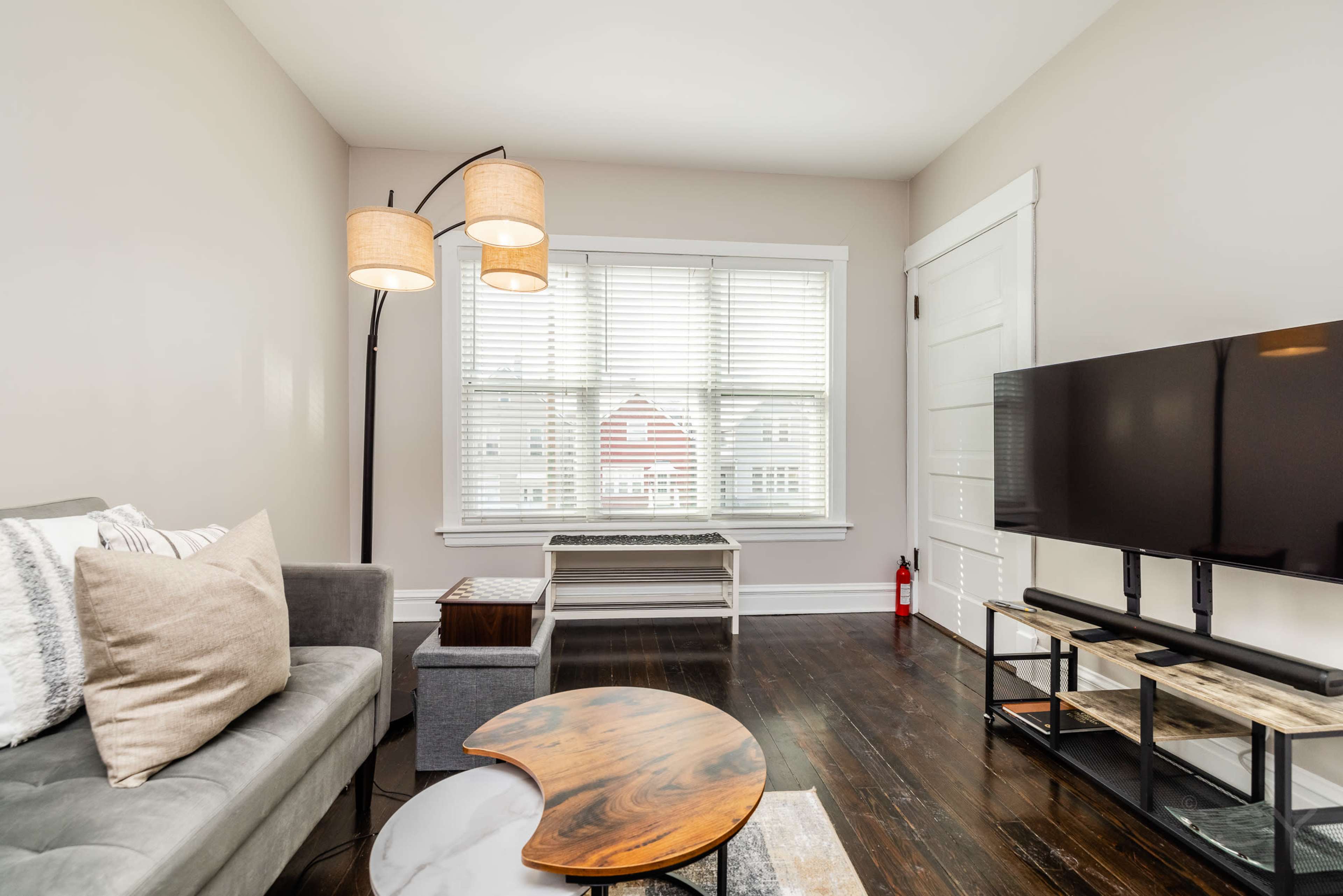 A living room features a gray sofa, a floor lamp with two shades, a coffee table, a television stand, and a window with white blinds.