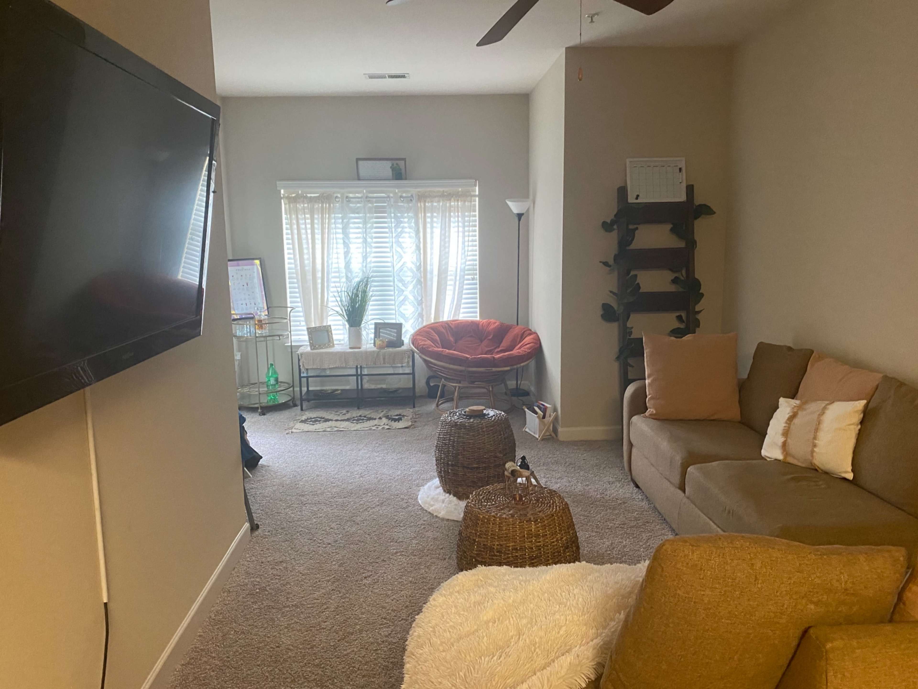 A living room with a large television, a sectional sofa, a round chair, and two woven ottomans.