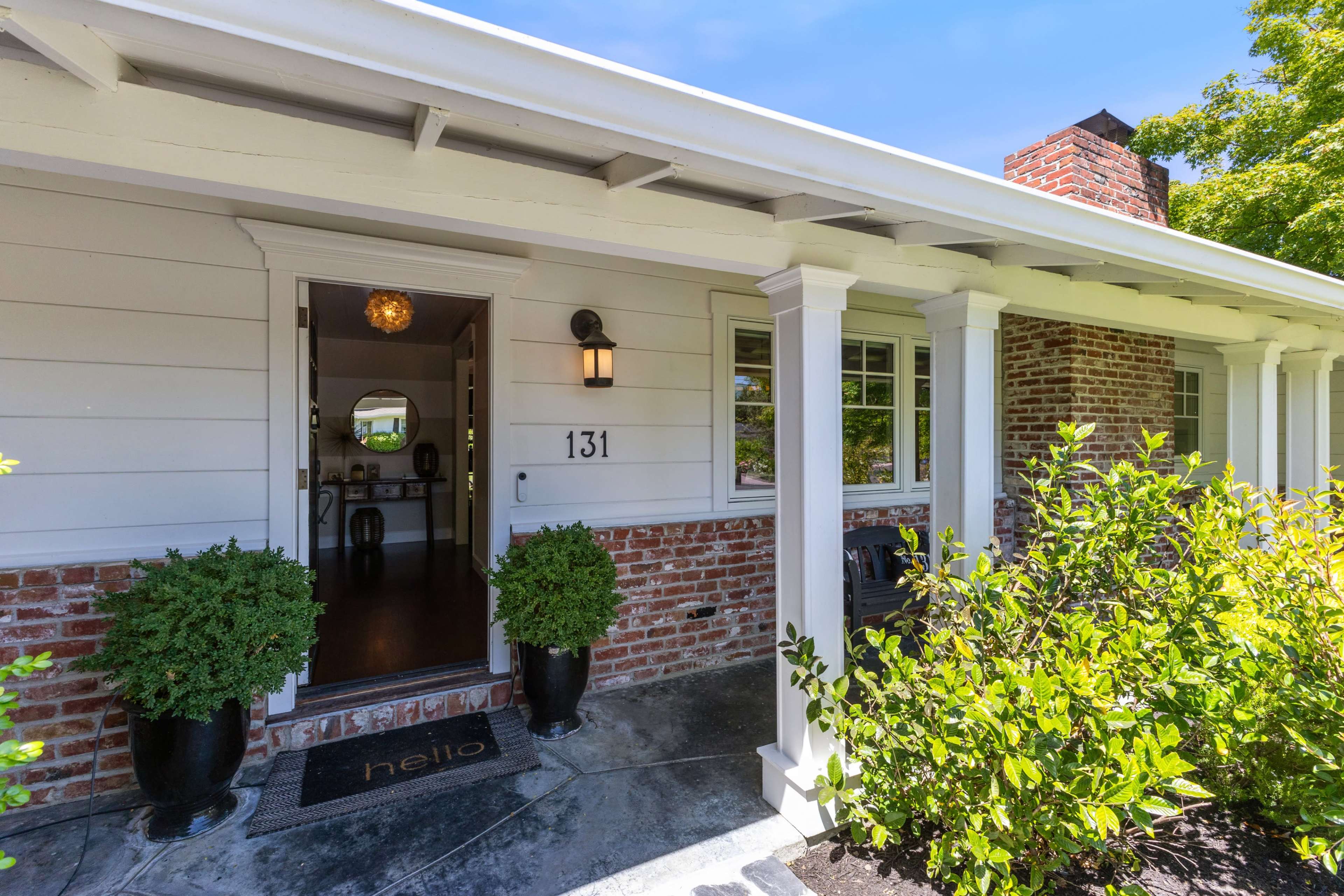 The entrance of a house features a front porch with a welcome mat, decorative plants, and a brick facade.