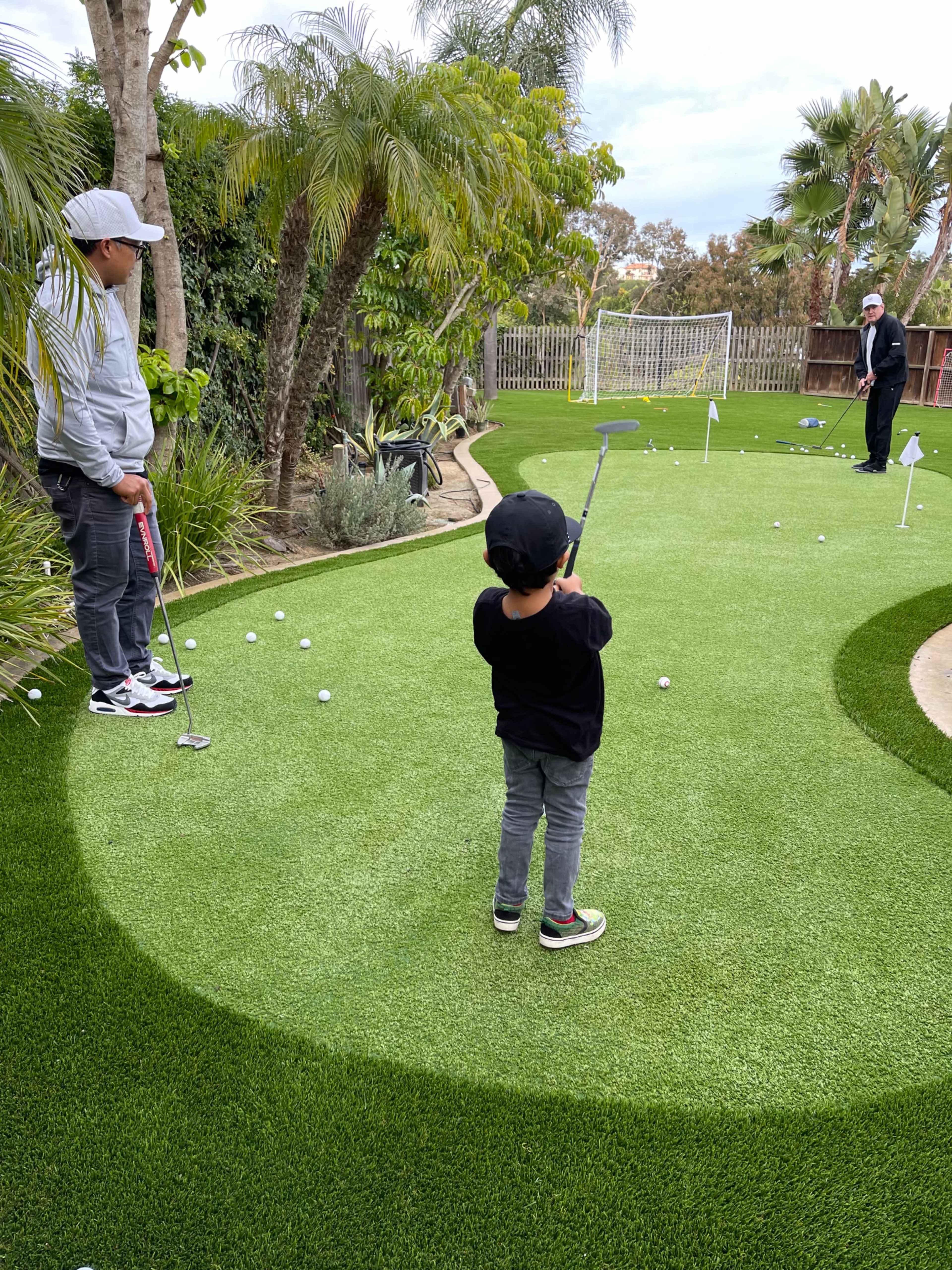 A child practices putting on a mini-golf green in a backyard while an adult observes.