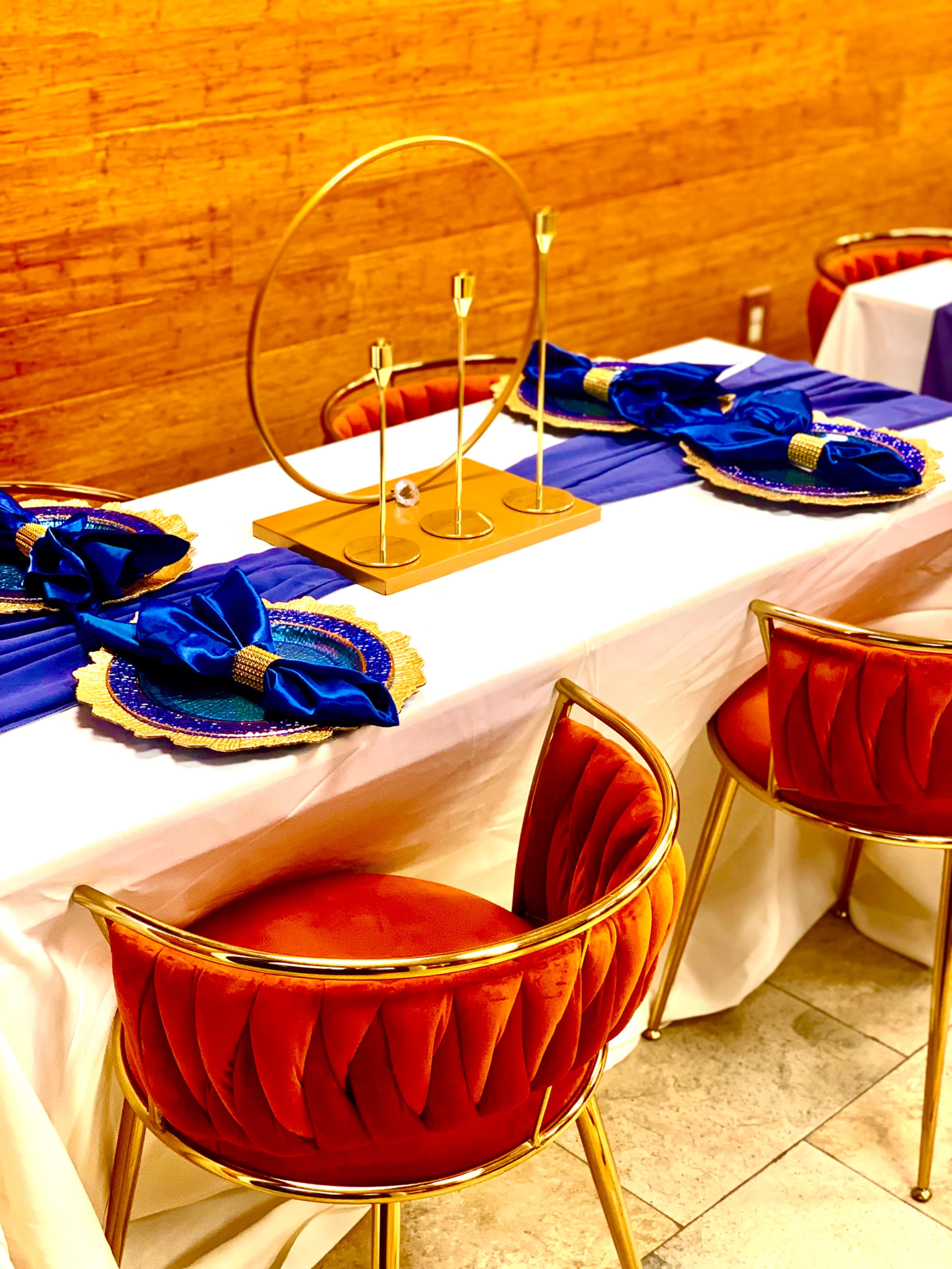 A neatly arranged dining table with blue napkins, gold-accented plates, and stylish golden centerpieces placed on a white tablecloth.