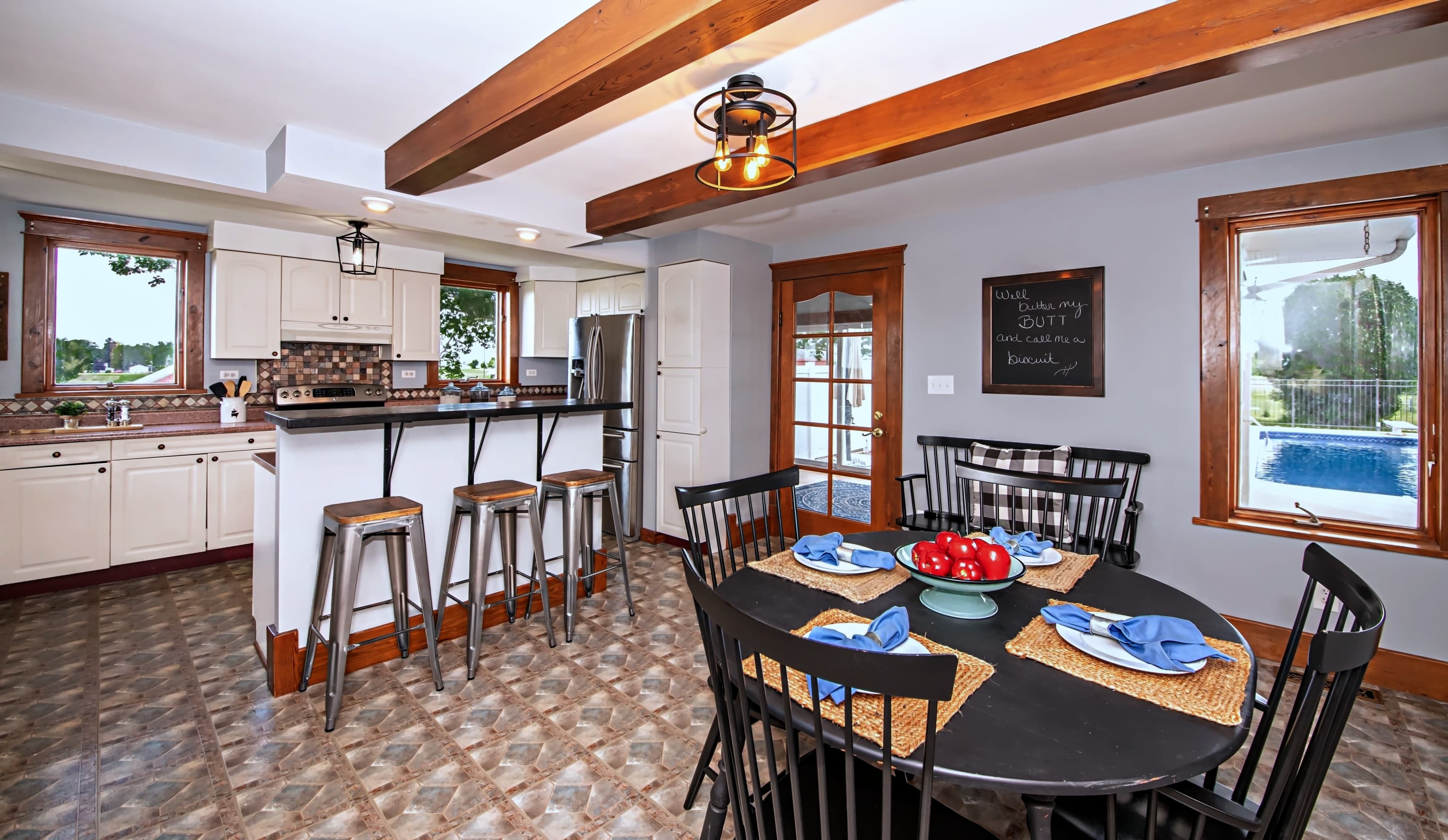 A modern kitchen features a dining area with a black table, bar seating, and a view of a pool outside.