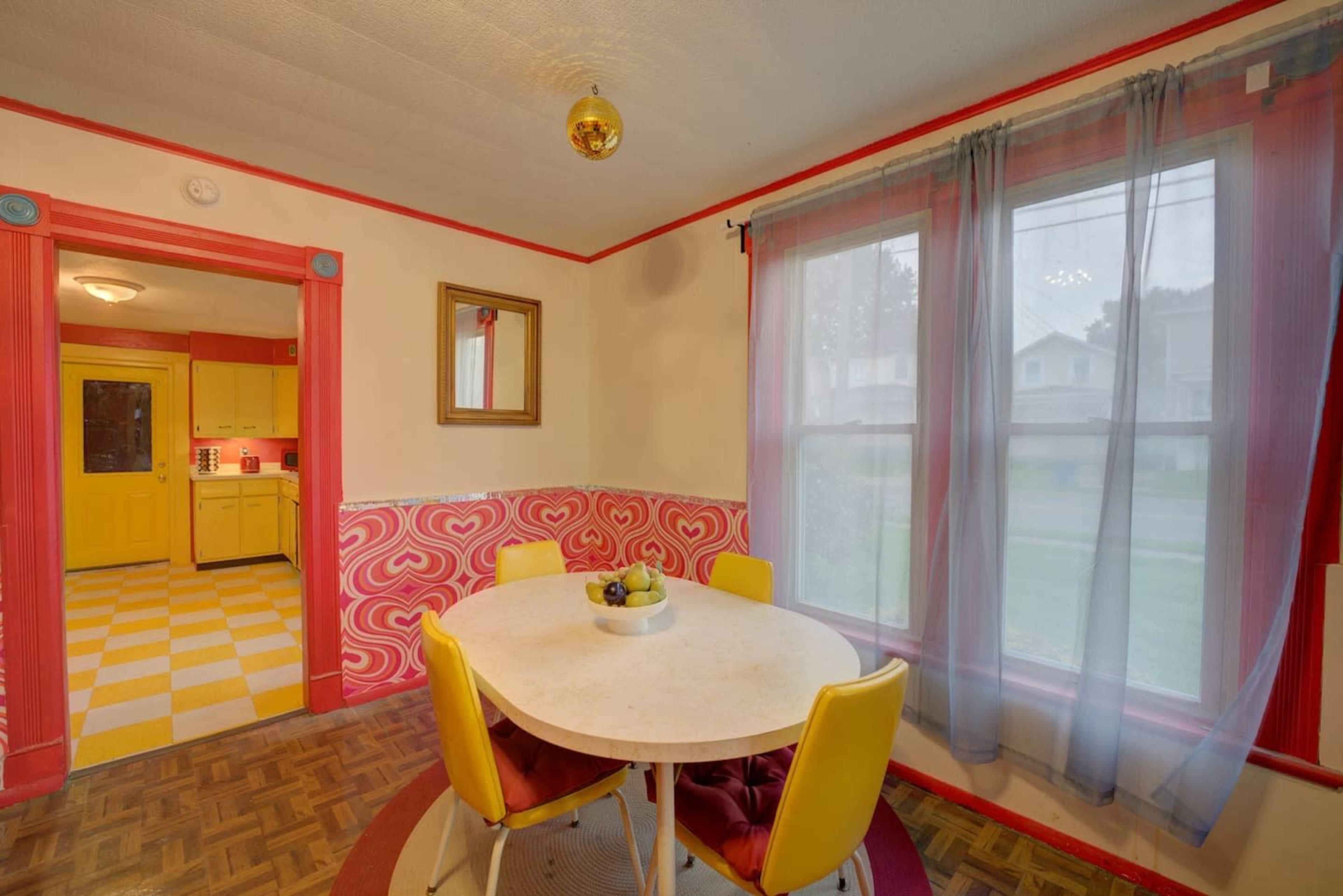 A dining area features a round table surrounded by yellow chairs, with red and white patterned wallpaper and a large window draped with sheer curtains.