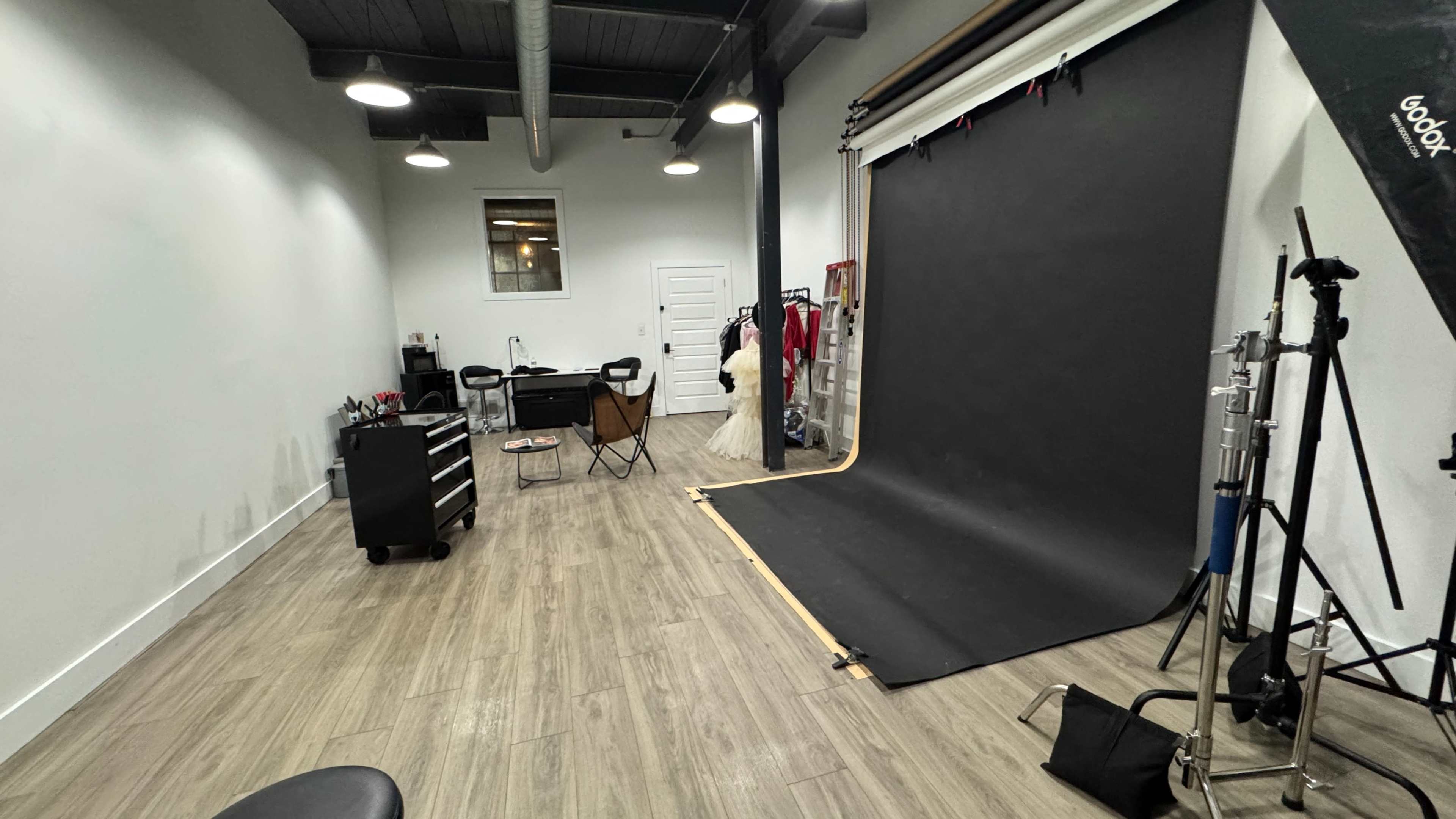 The image shows a photography studio with a black backdrop, a makeup station, a chair, and various photography equipment arranged along the walls.