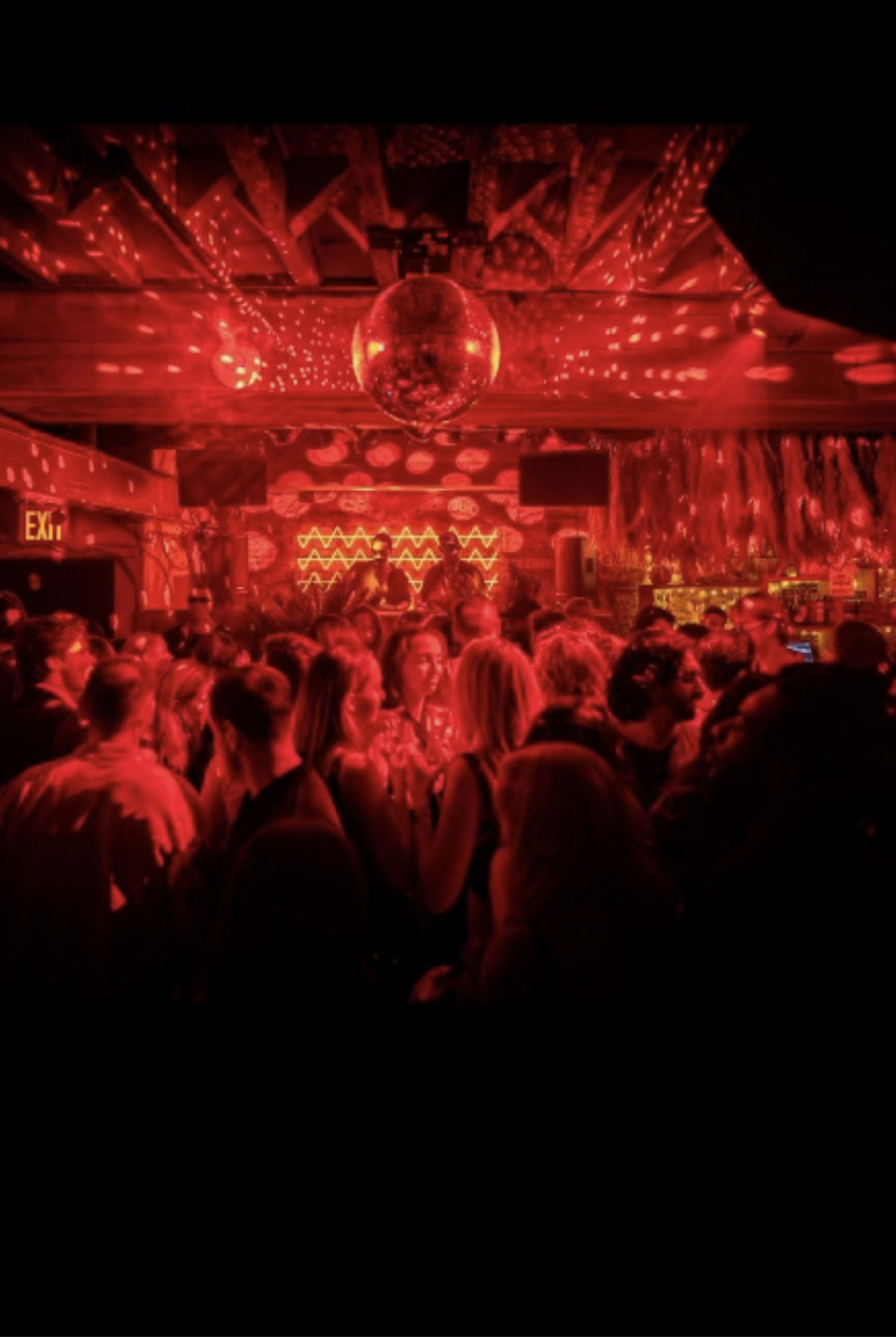 A crowded nightclub features red lighting and a disco ball, creating a lively atmosphere.