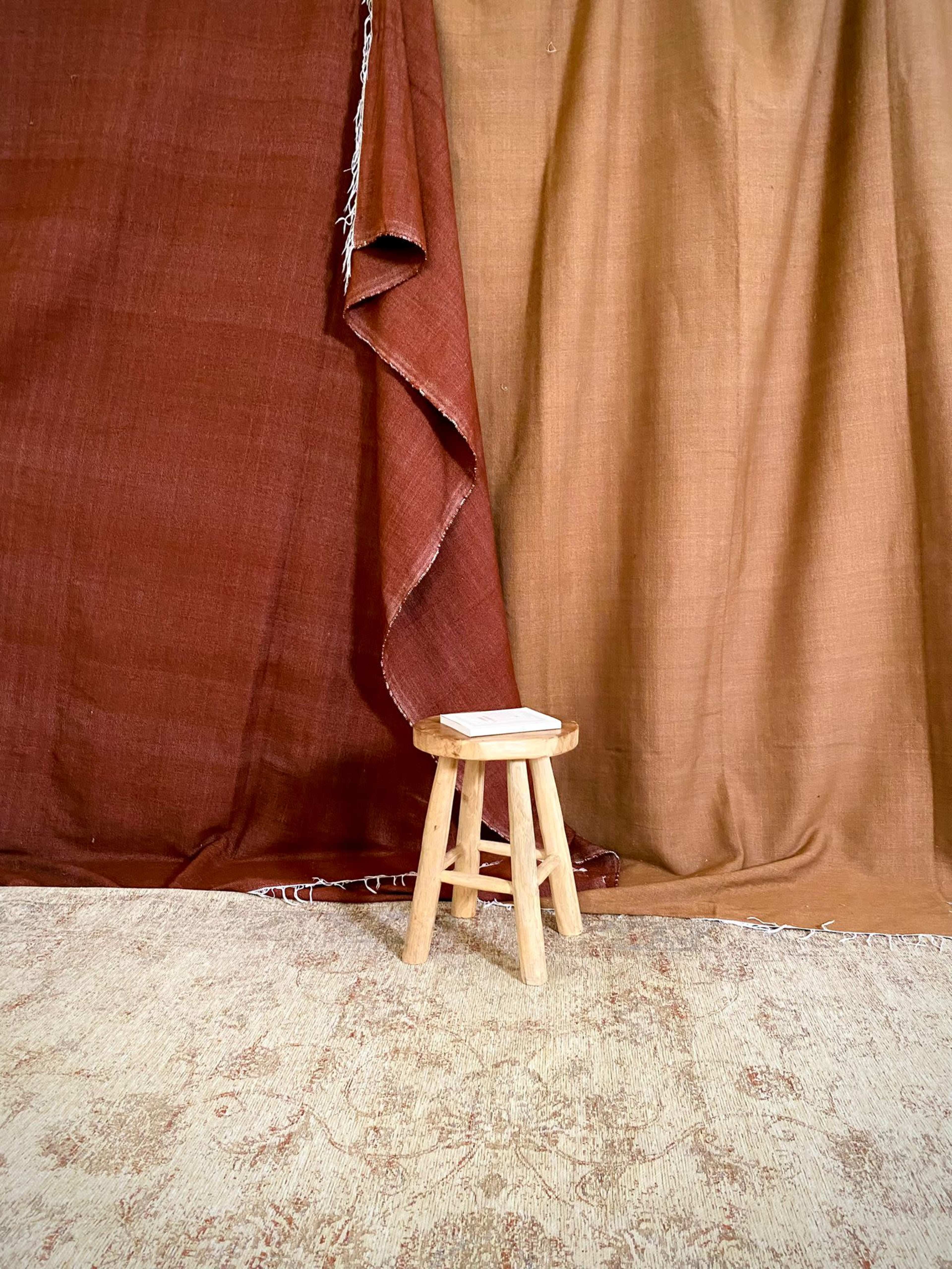A small wooden stool stands on a patterned rug in front of two draped fabric backdrops in earthy tones.