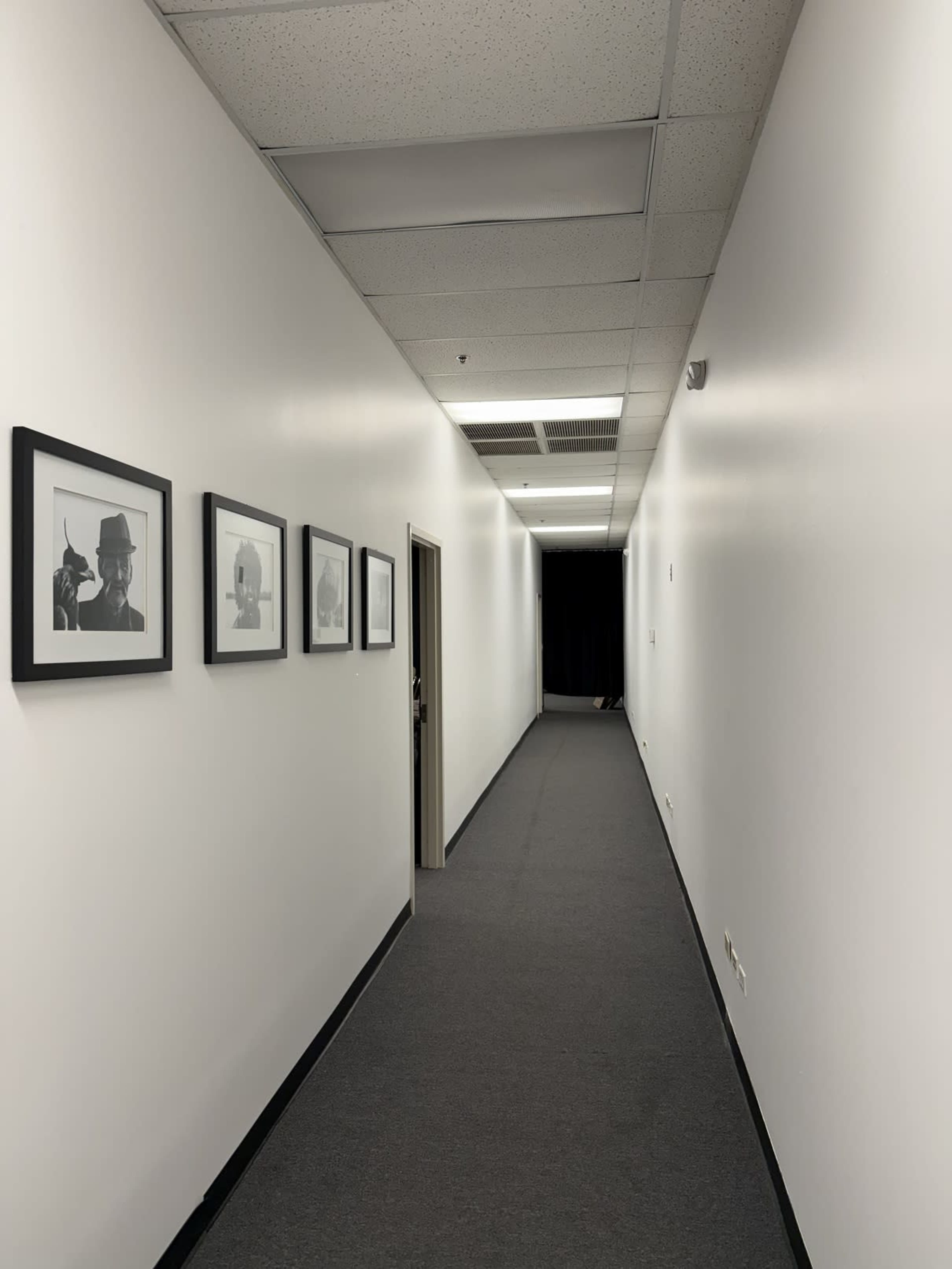 A narrow hallway with white walls and a gray carpet features several framed black-and-white photographs on one side.