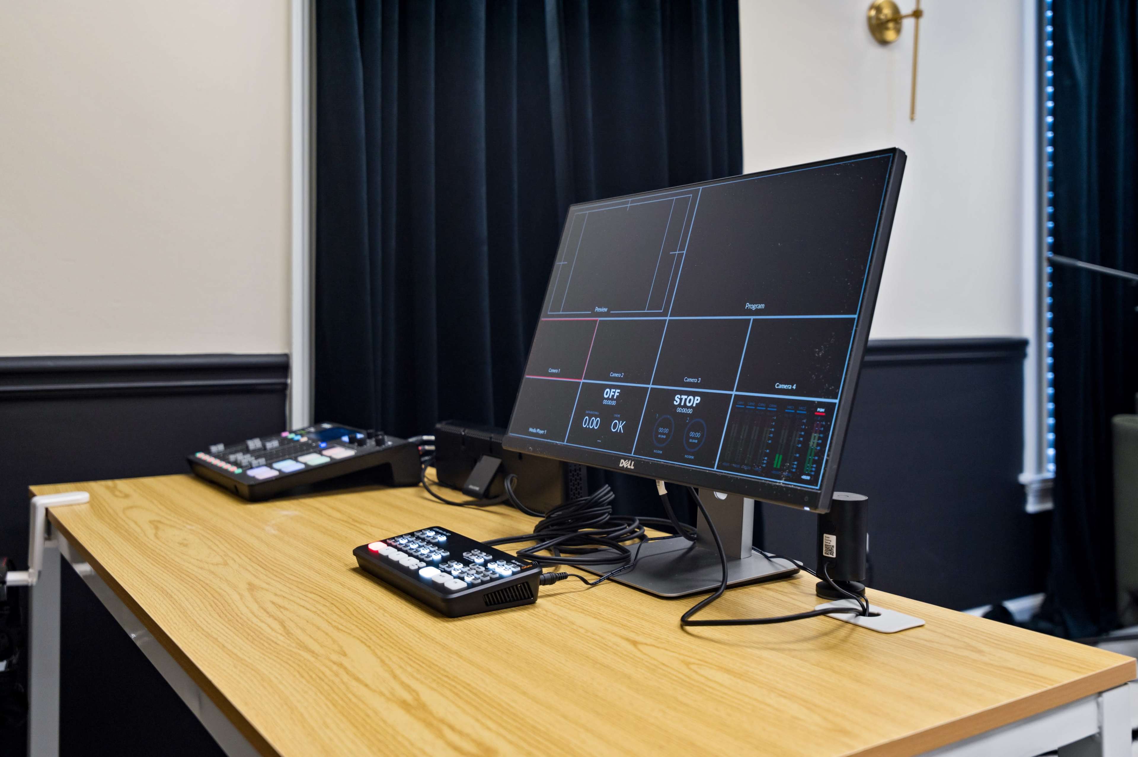 A computer monitor displaying multiple input feeds is positioned on a wooden desk alongside a control panel with buttons and a digital display.