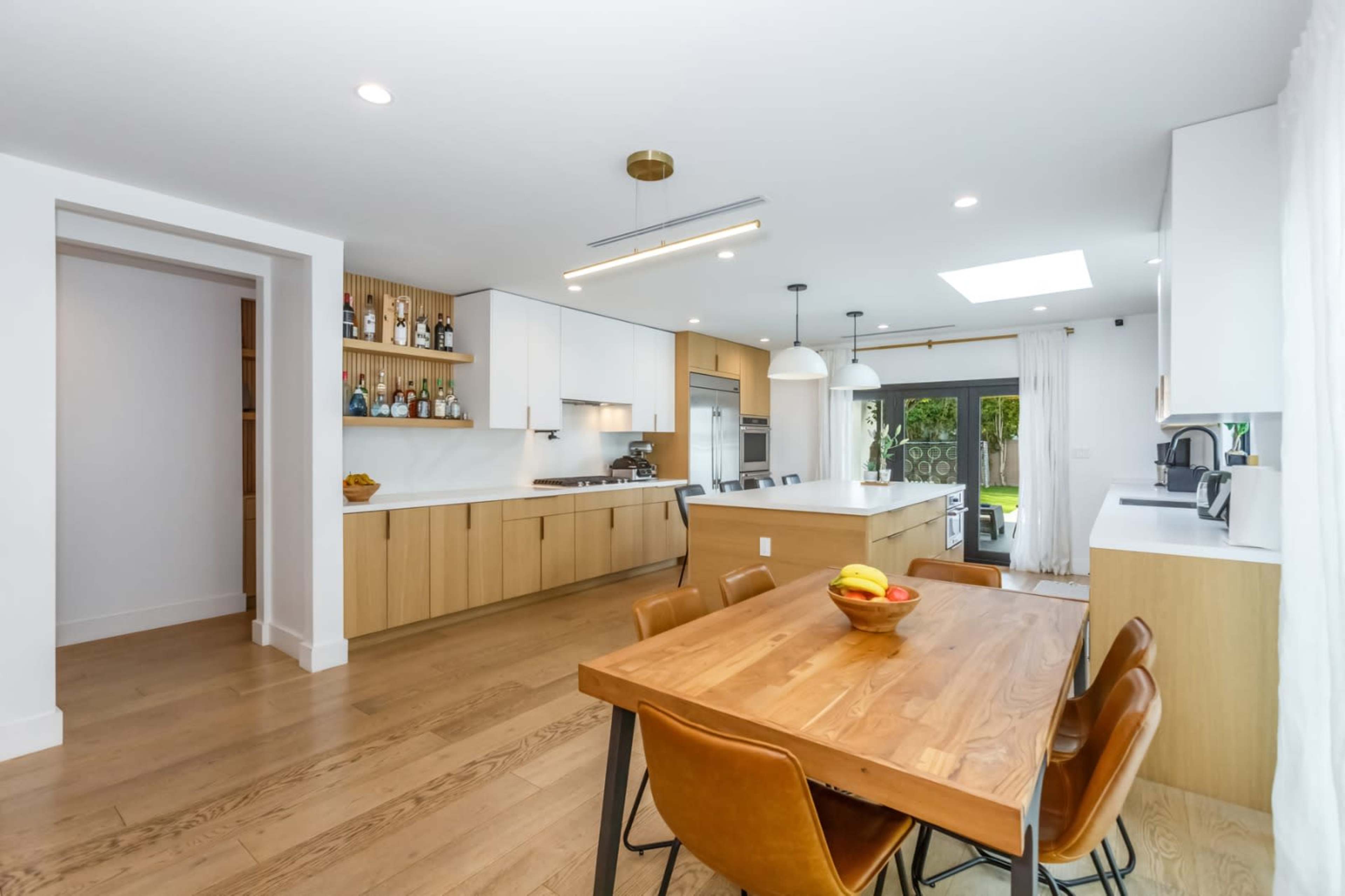 A modern kitchen features light wooden cabinetry, a large wooden dining table, and an abundance of natural light from a ceiling skylight.
