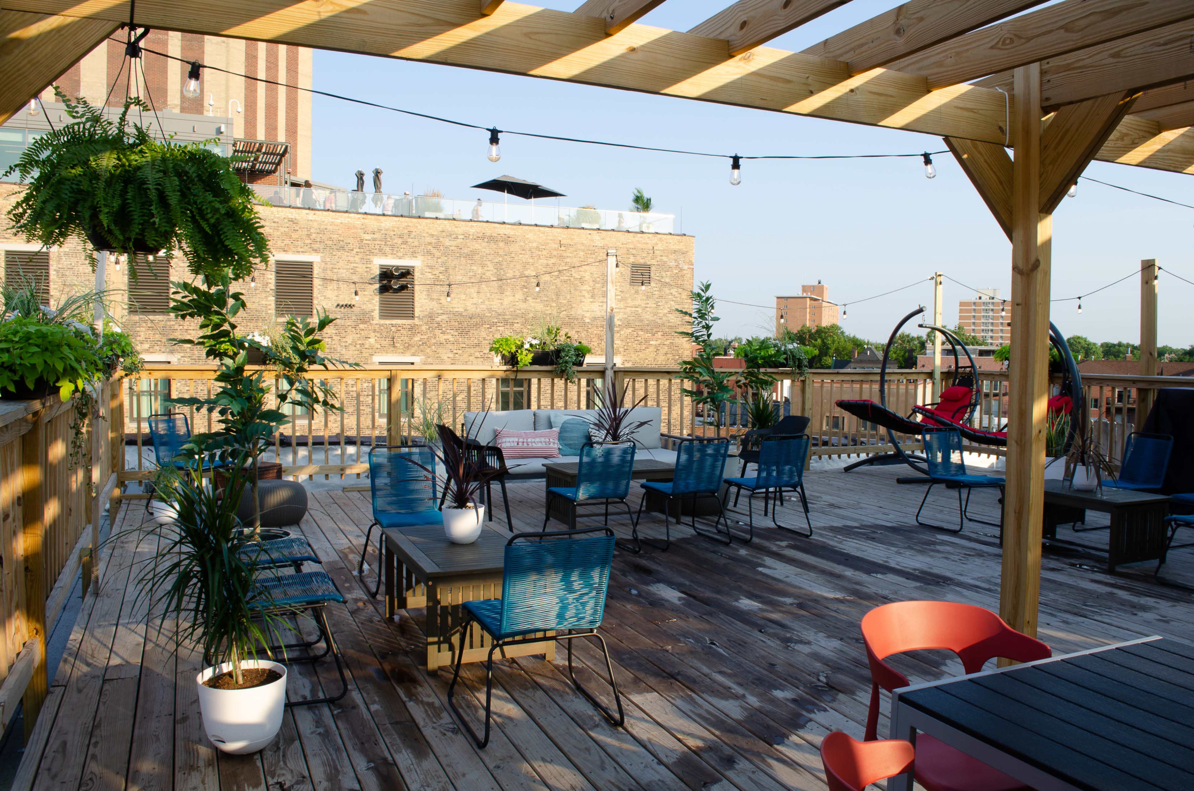 A rooftop patio features wooden decking, multiple seating areas with tables and chairs, and hanging plants under a wooden pergola.