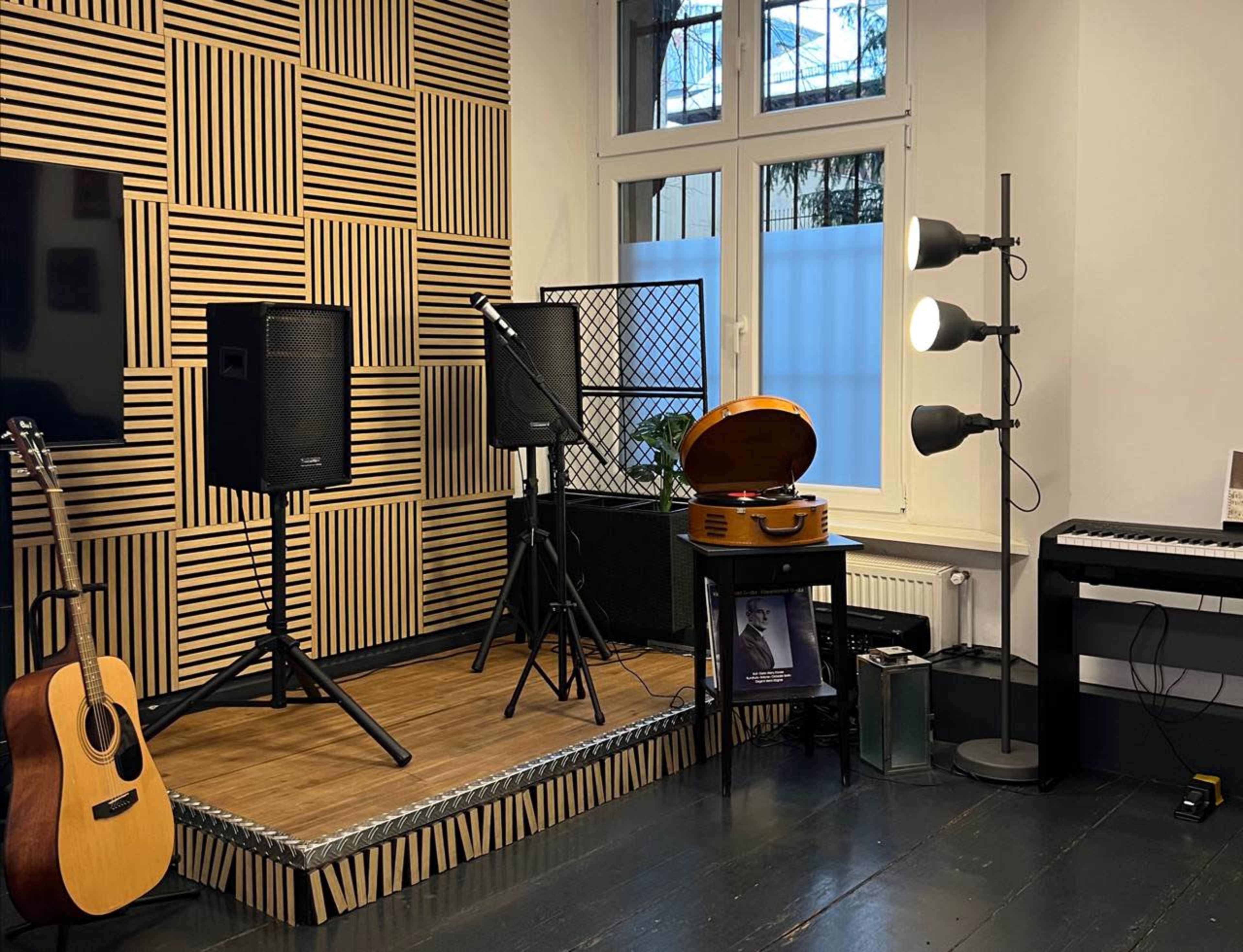 The image shows a music studio setup featuring two speakers, a vintage record player, an acoustic guitar, a keyboard, and a standing lamp against a wall with a patterned wooden design.