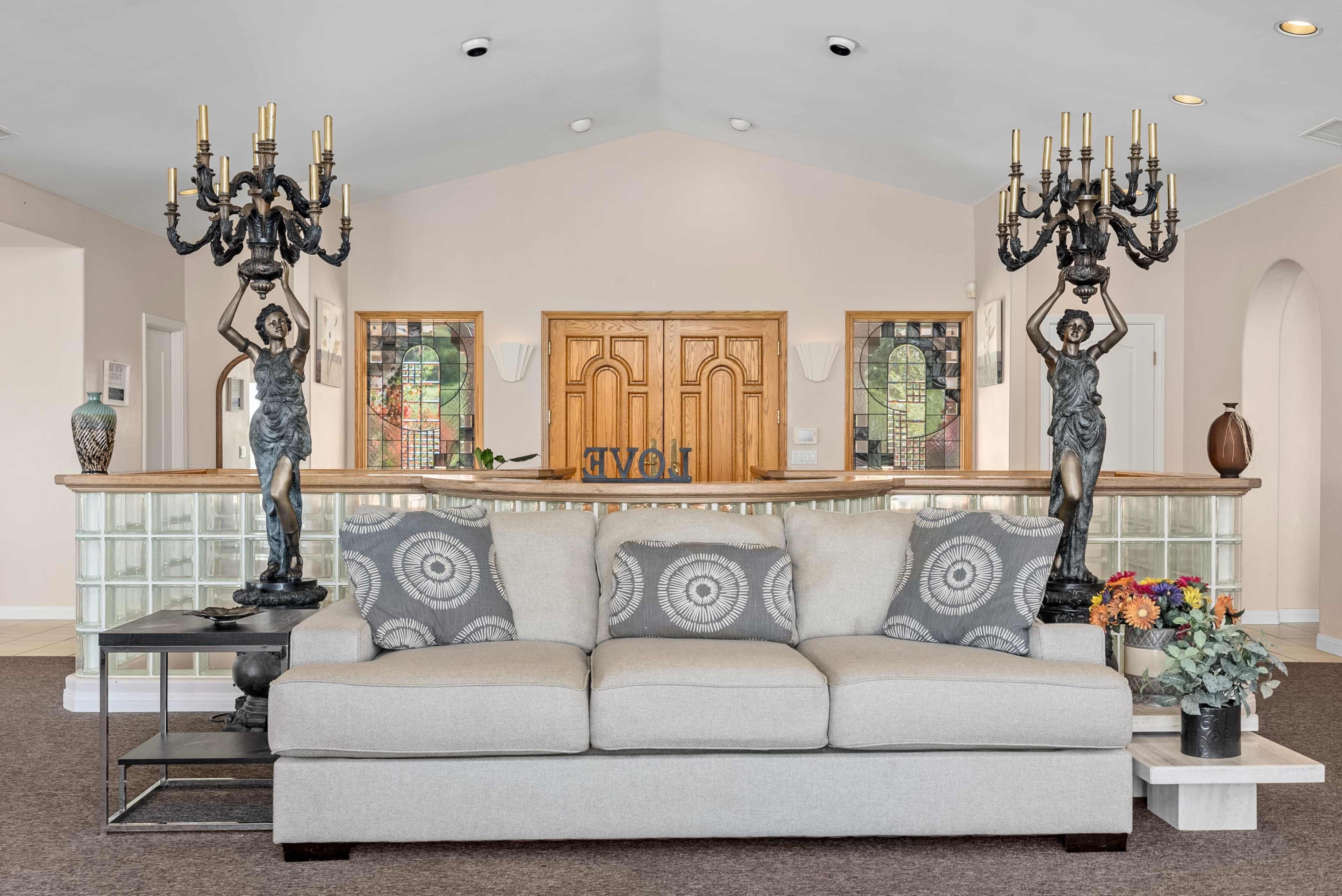 A living area with a light gray sofa adorned with round patterned pillows, flanked by two tall candelabra-style lamps and a wooden reception desk featuring stained glass windows in the background.