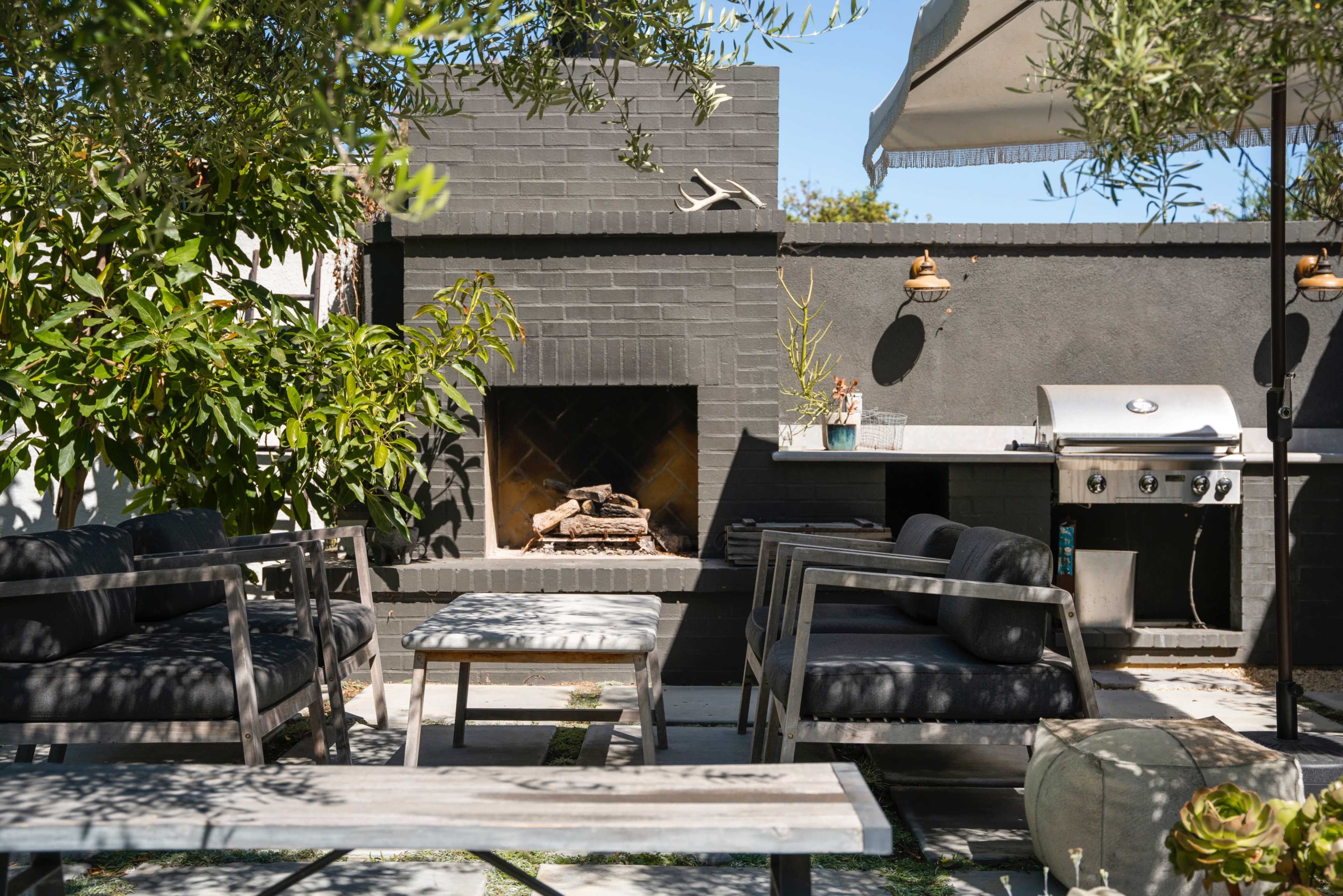 The image shows an outdoor seating area with a fireplace, a grill station, and shaded seating surrounded by greenery.