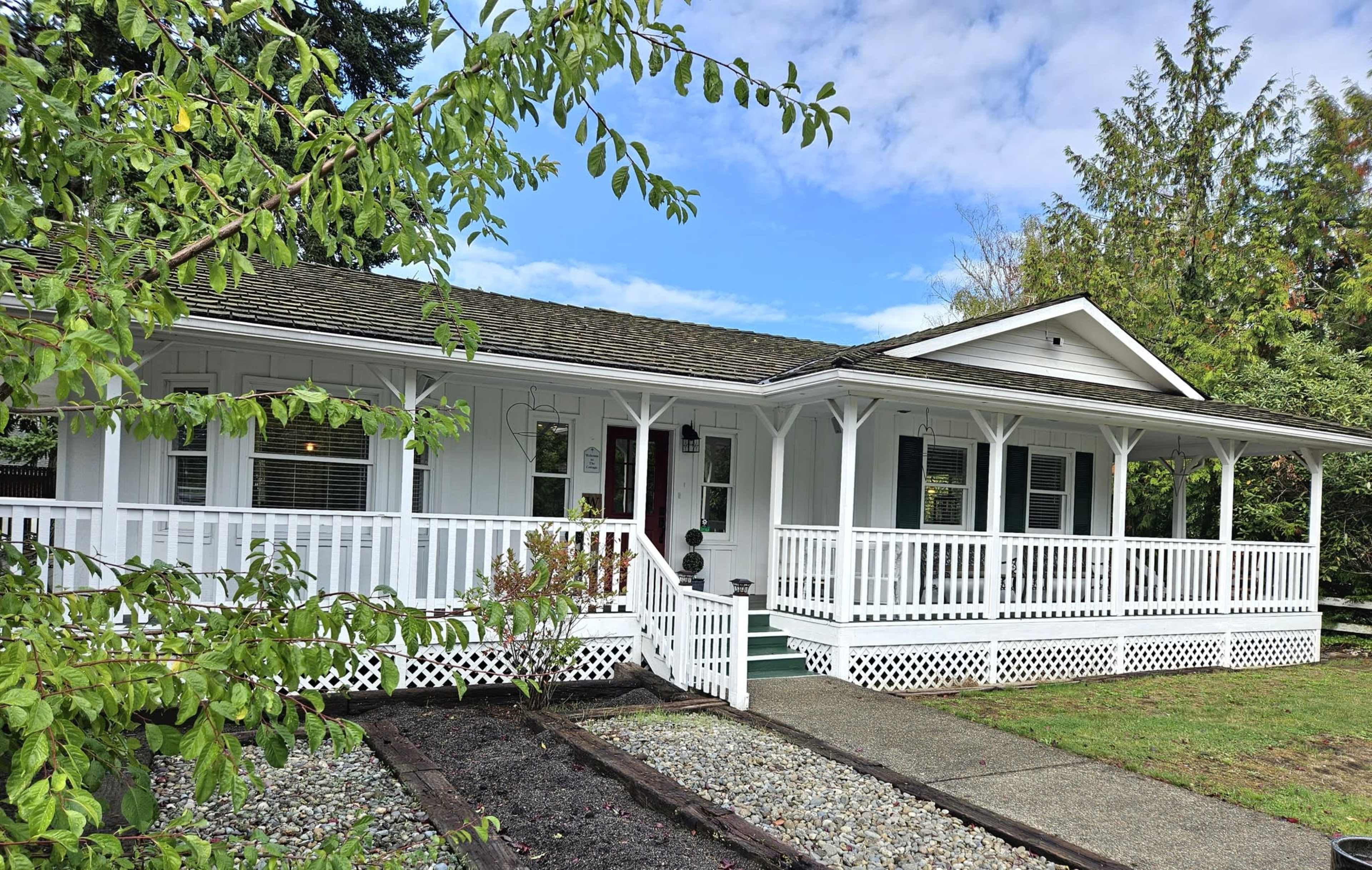 The image shows a white, single-story house with a wide porch, surrounded by greenery and a gravel walkway.
