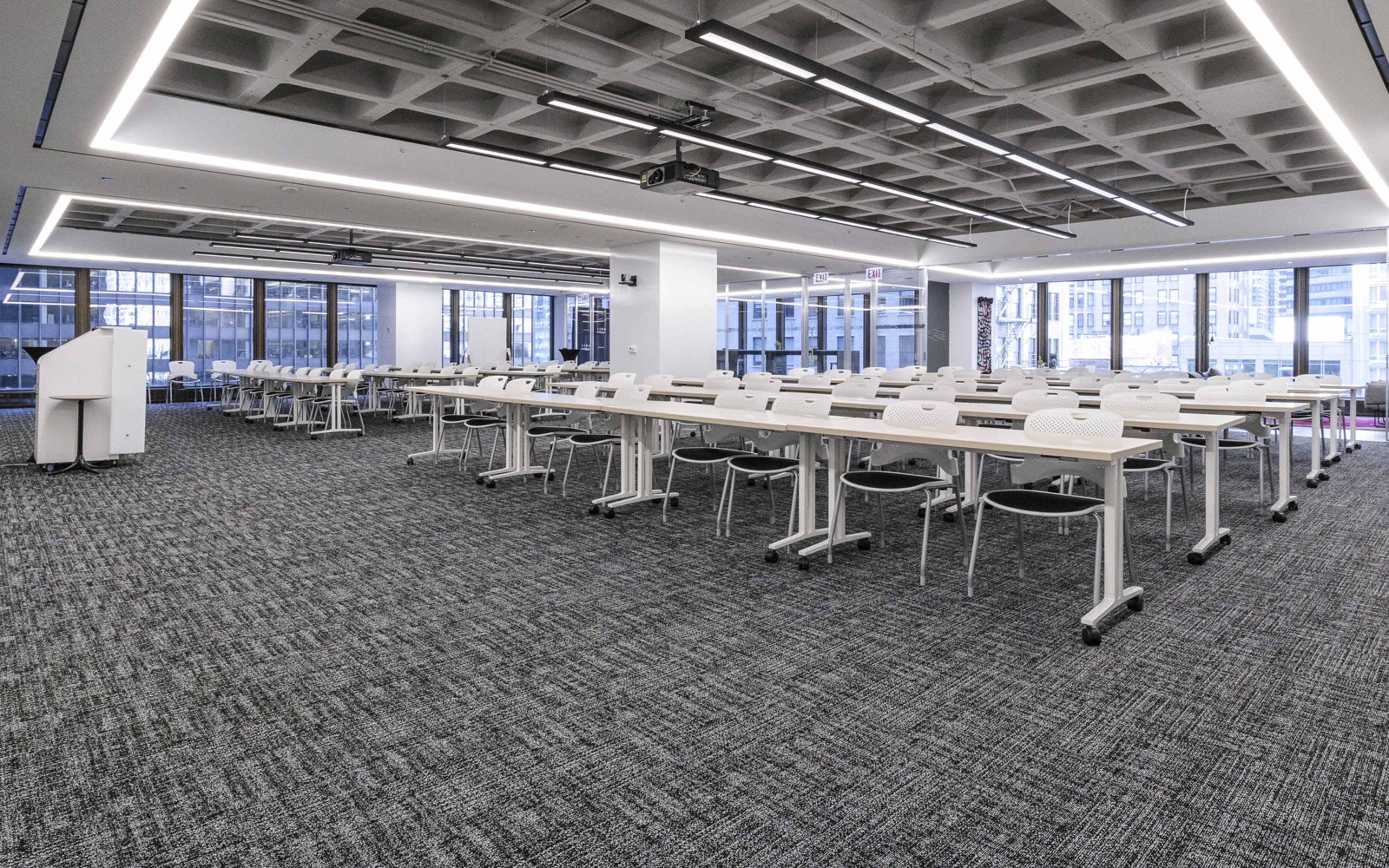 A spacious, modern classroom is arranged with multiple white tables and chairs, featuring large windows along one wall that overlook a cityscape.