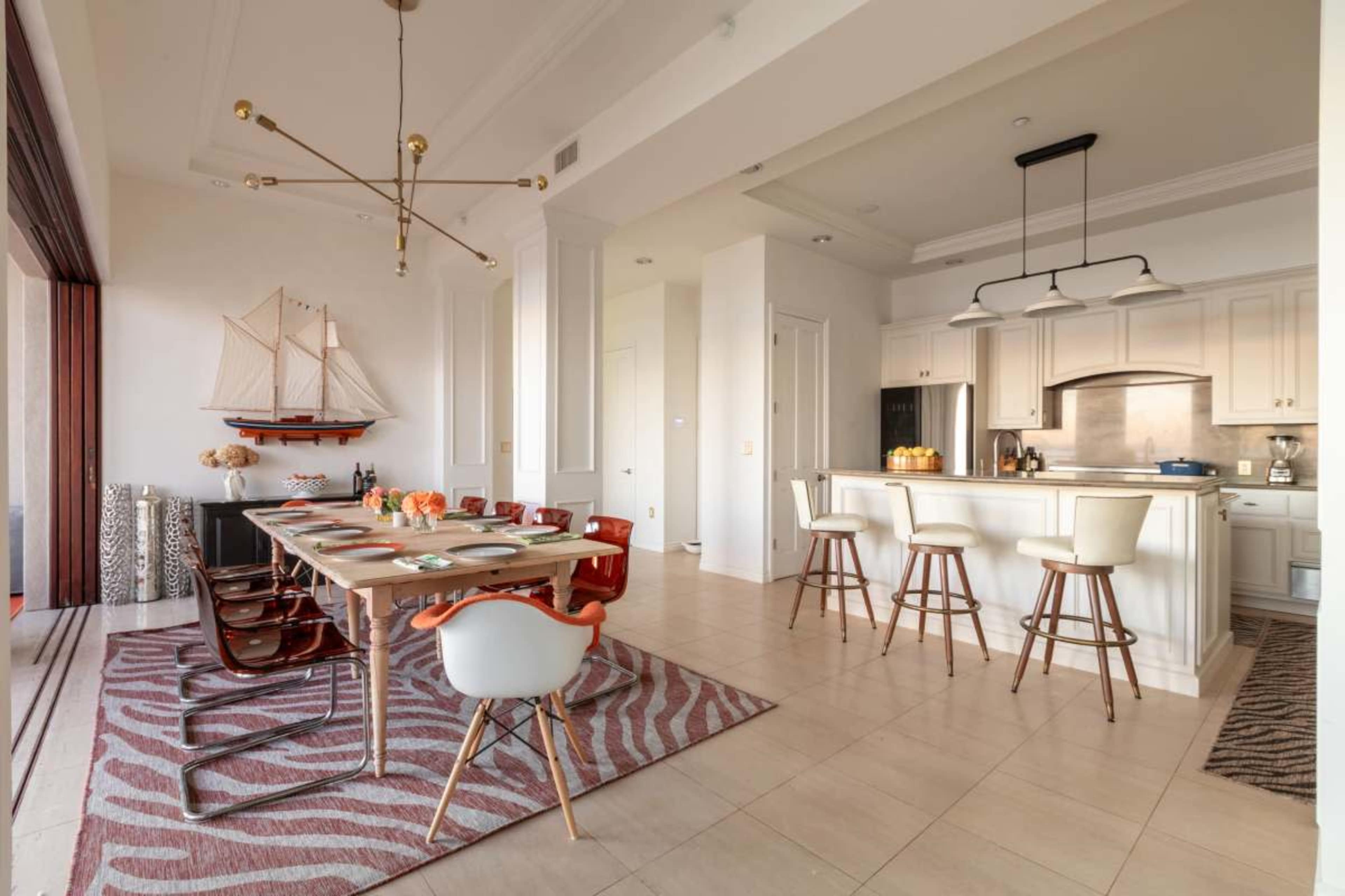 The image shows a modern dining area with a large wooden table set for a meal, surrounded by various chairs, and a kitchen counter with bar stools in an open-concept space.