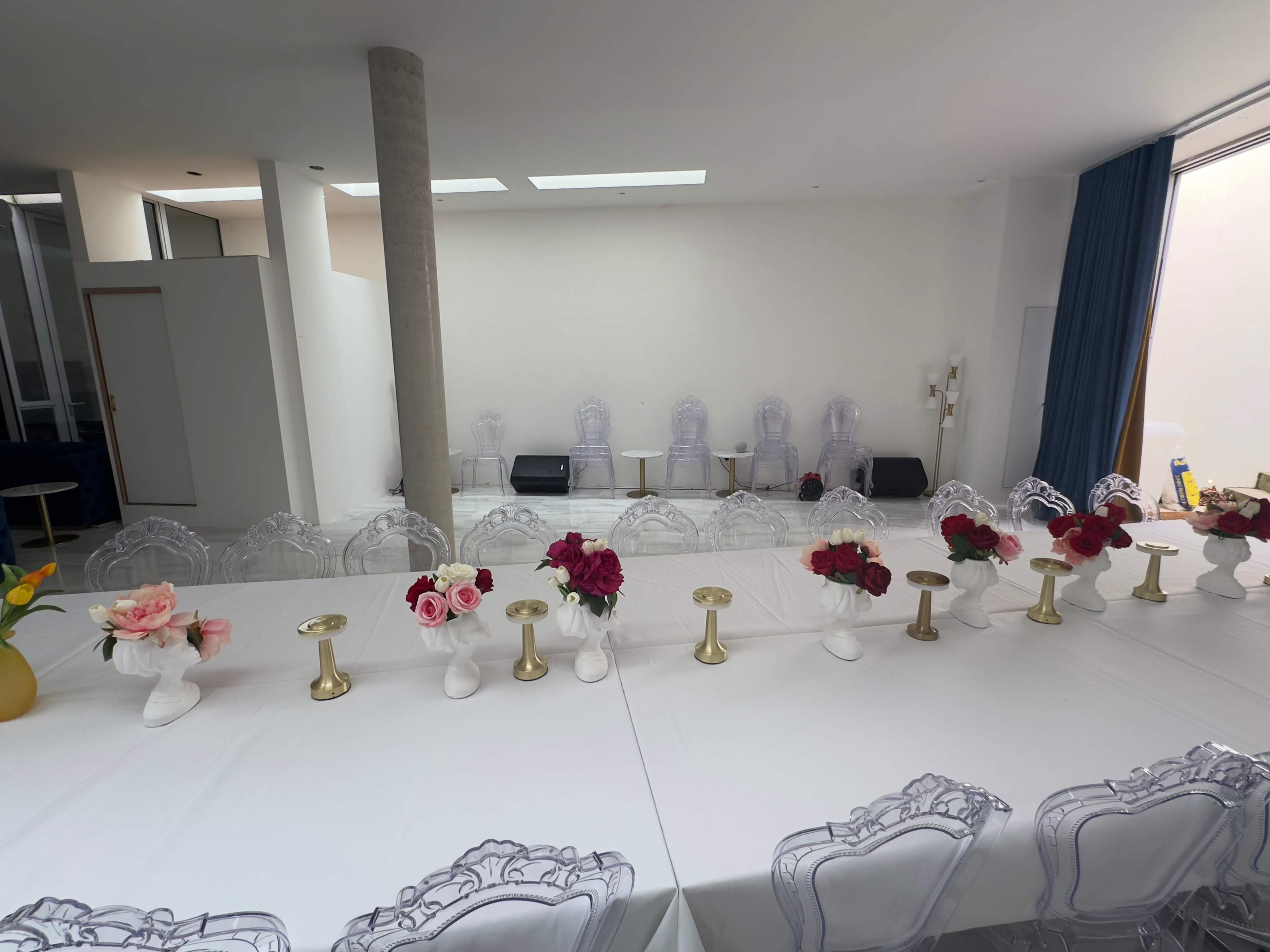 A long dining table is set in a bright room with transparent chairs, floral centerpieces, and gold candlesticks.