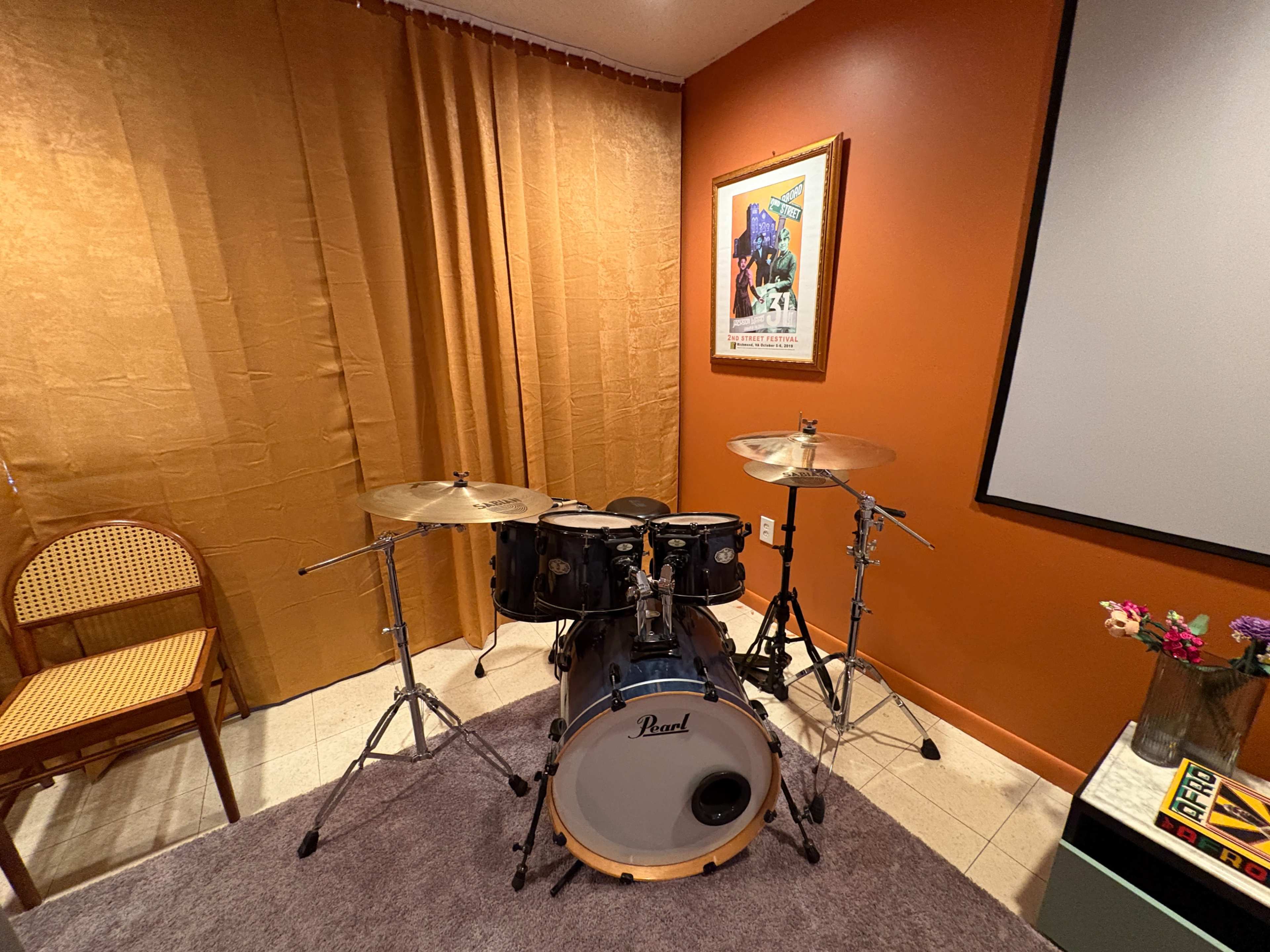 A black drum set is positioned in a room with orange walls and a large curtain, near a chair and a framed artwork.