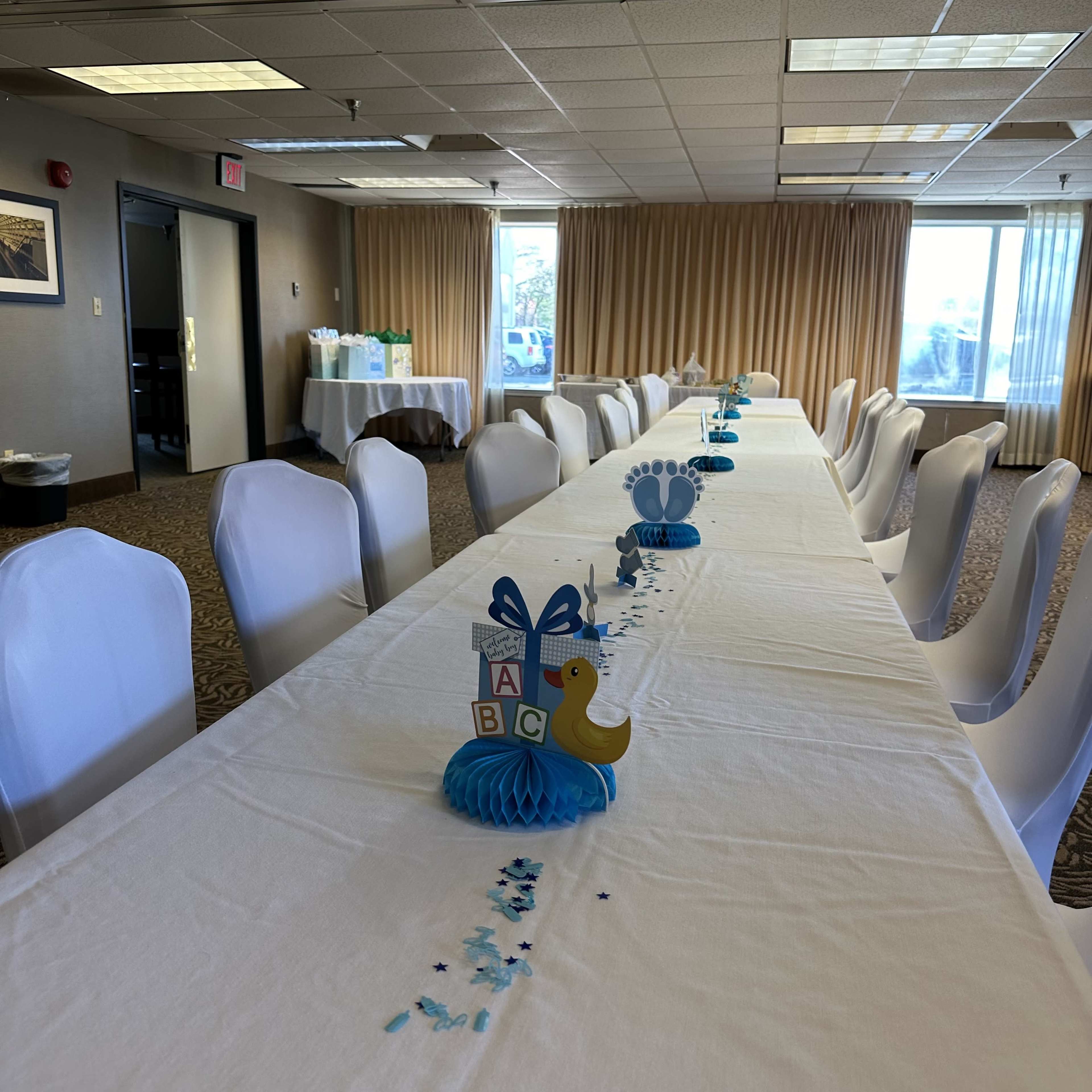 A long, decorated table with white chairs is set up in a room, featuring blue and yellow centerpieces and scattered confetti.