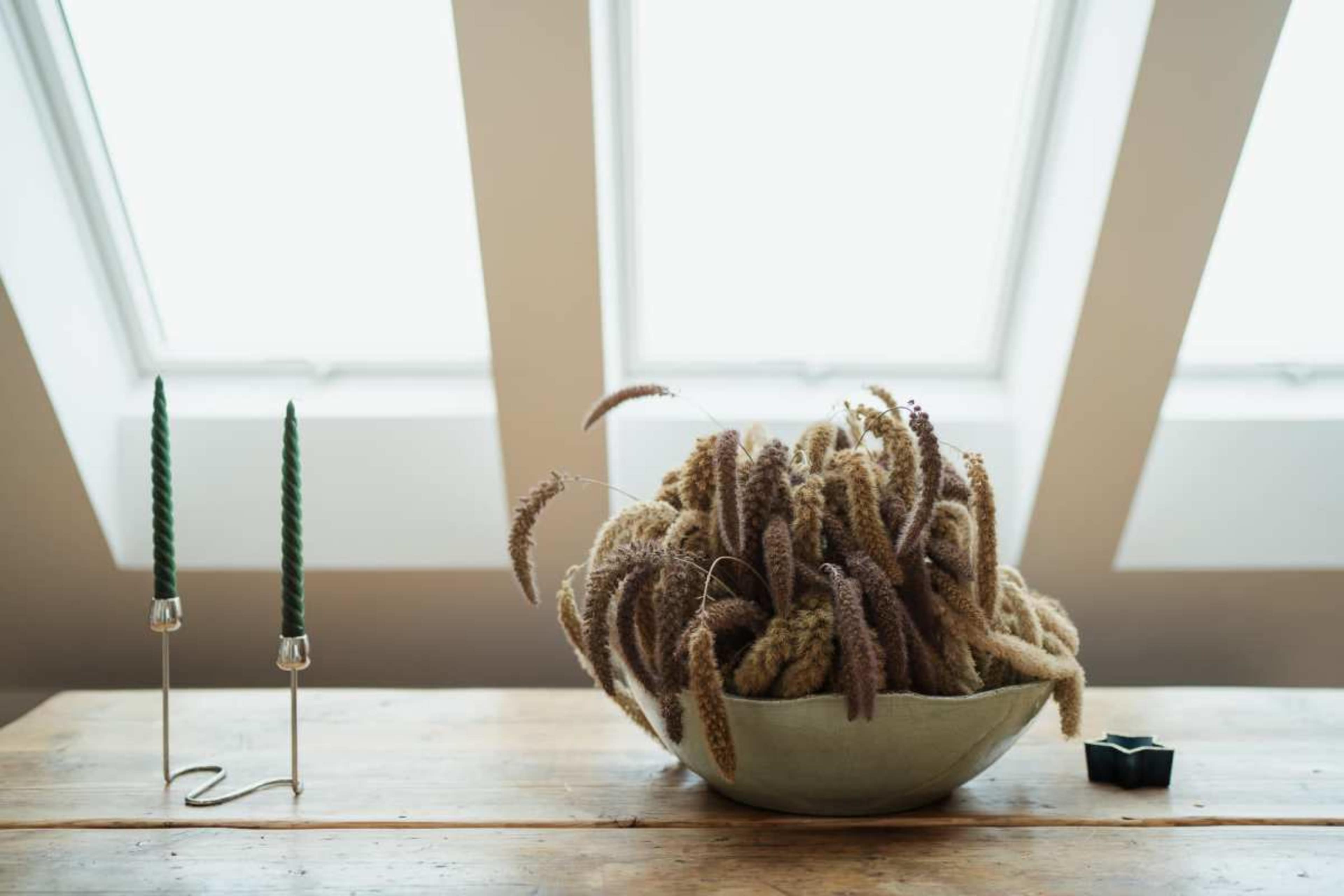 A light-filled room features a bowl of decorative dried plants on a wooden table, accompanied by two green candlesticks.