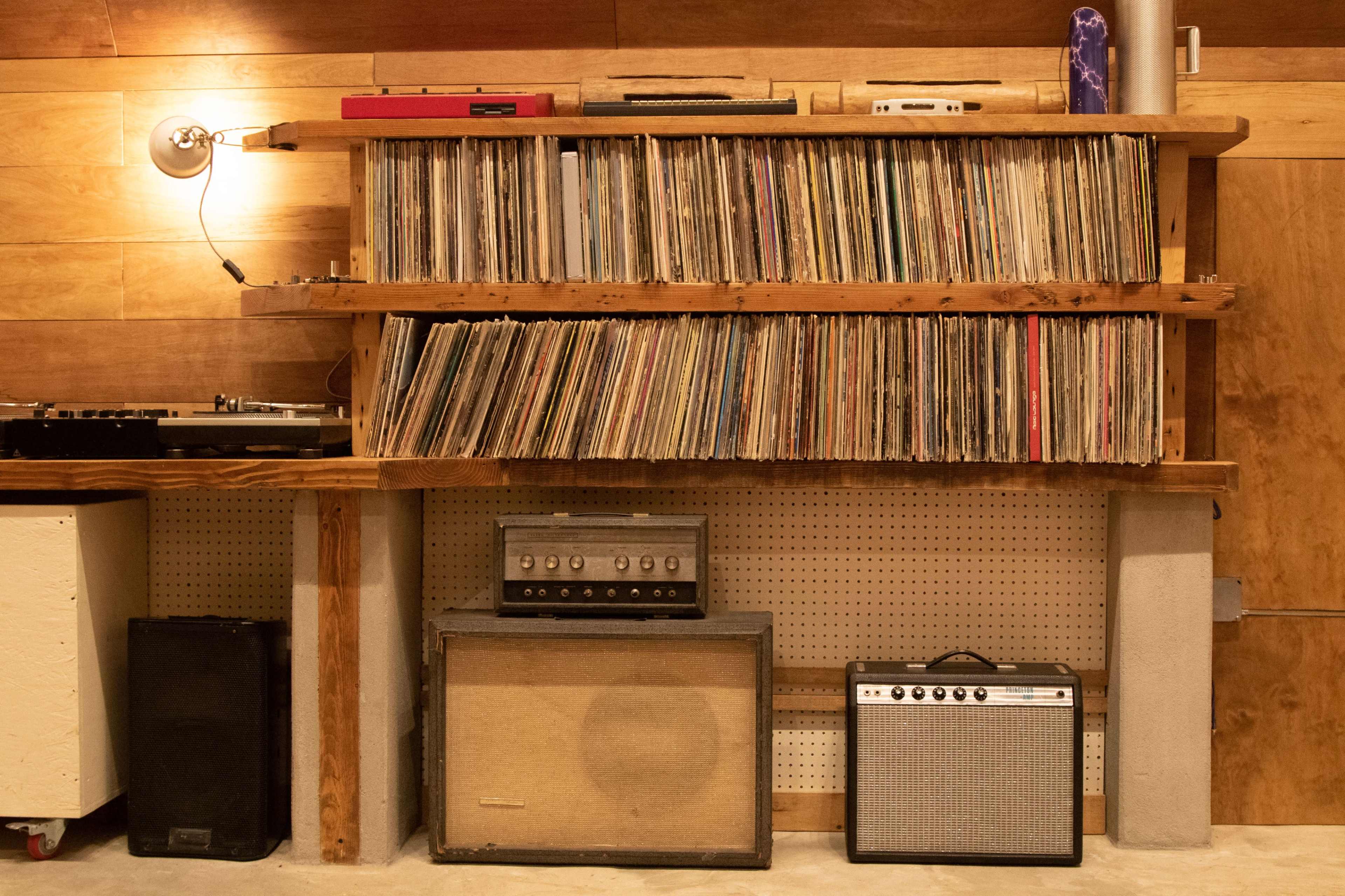 A wooden shelf holds a large collection of vinyl records above a stack of amplifiers and speakers.