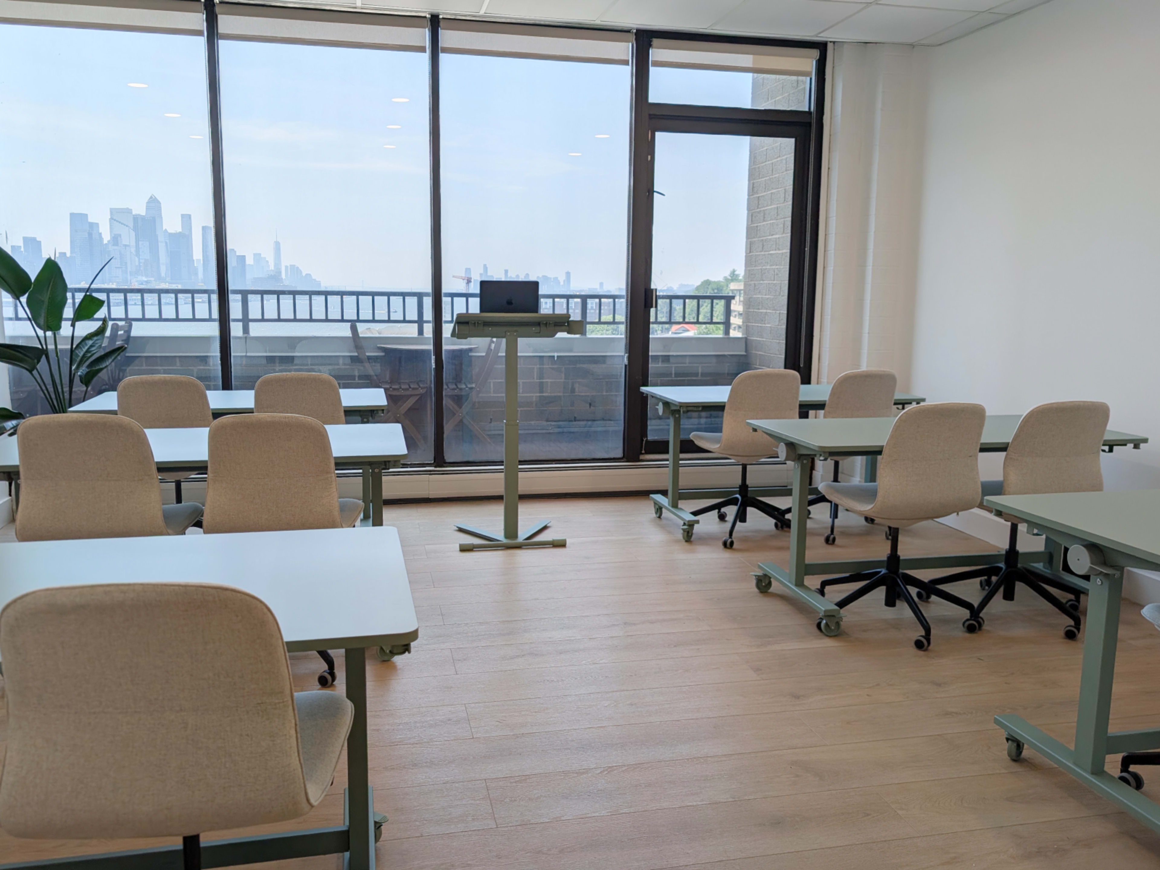 The image shows a bright classroom with several tables and chairs arranged neatly, featuring a large window that offers a view of a city skyline in the background.