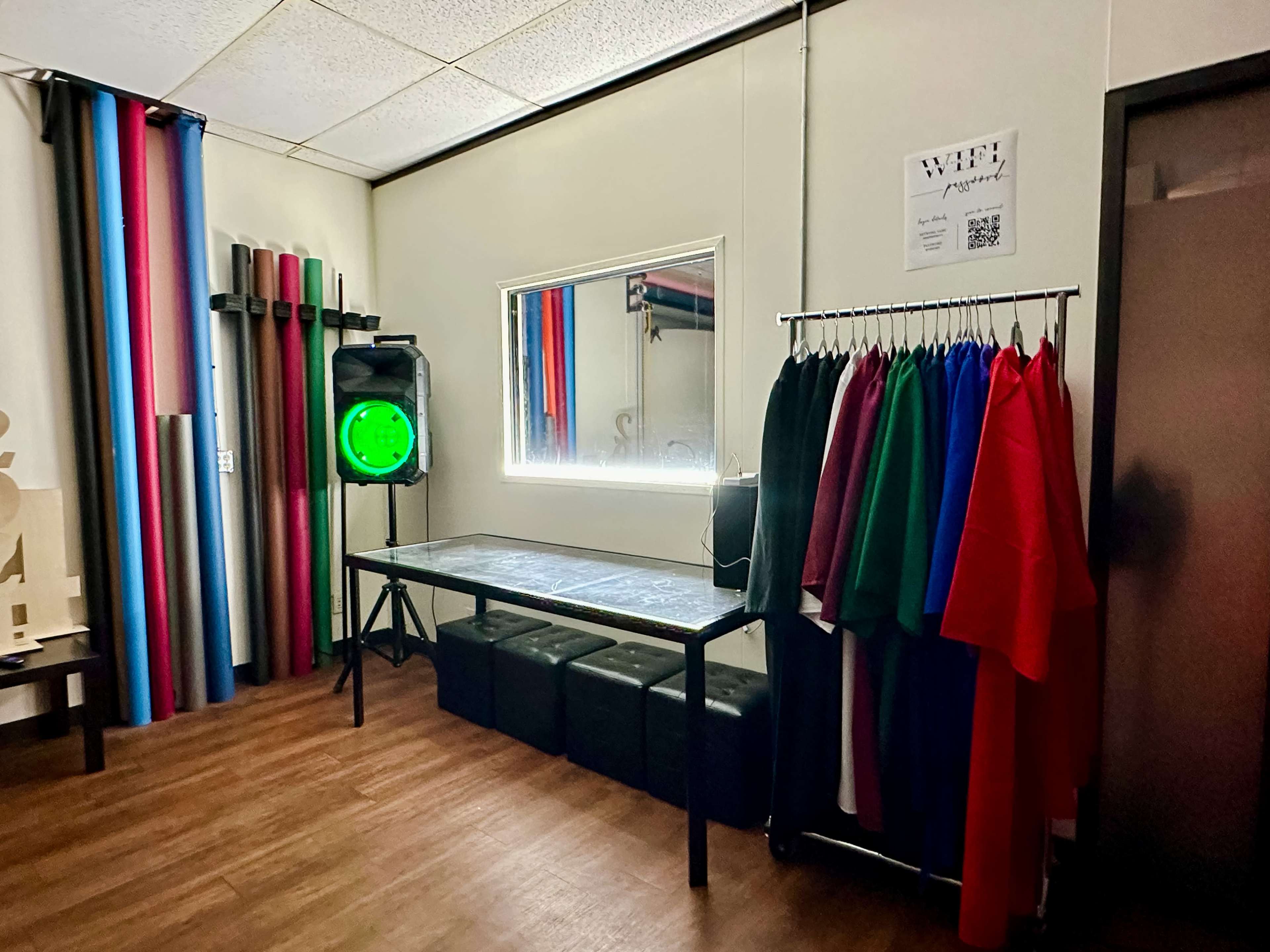 A room with a table, a green lighting fixture, a clothing rack filled with various colored garments, and colorful rolls of material along the wall.