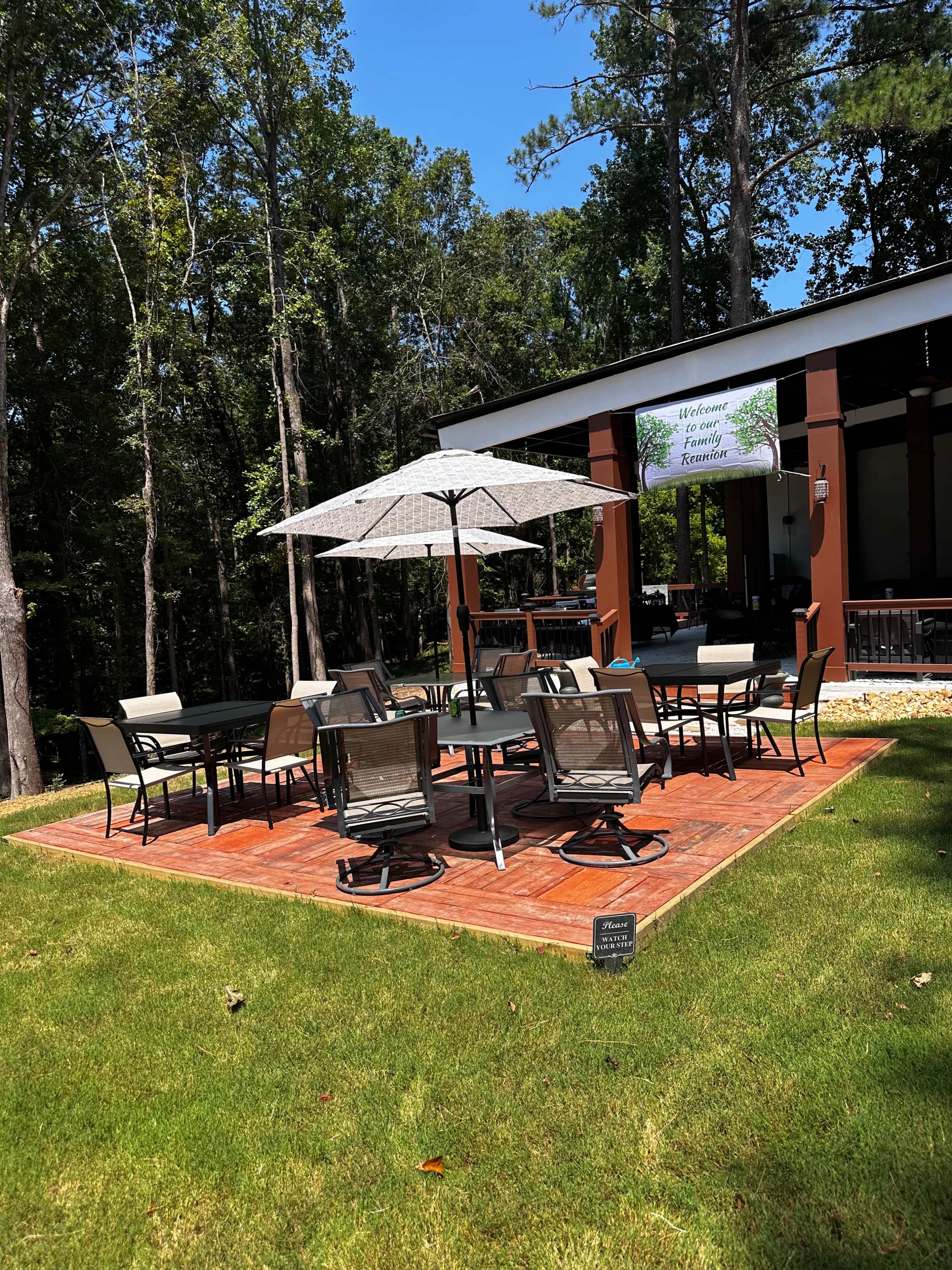 A patio area with a large umbrella, several dining tables and chairs, is situated on a grassy lawn surrounded by trees.
